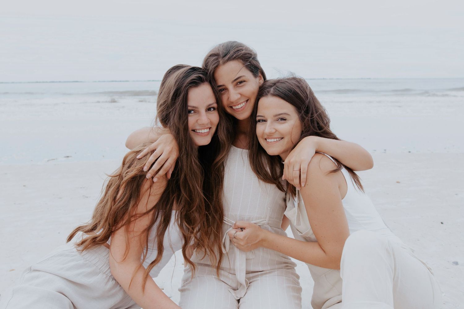 Three sisters in white dresses sharing joyful moment on beach.