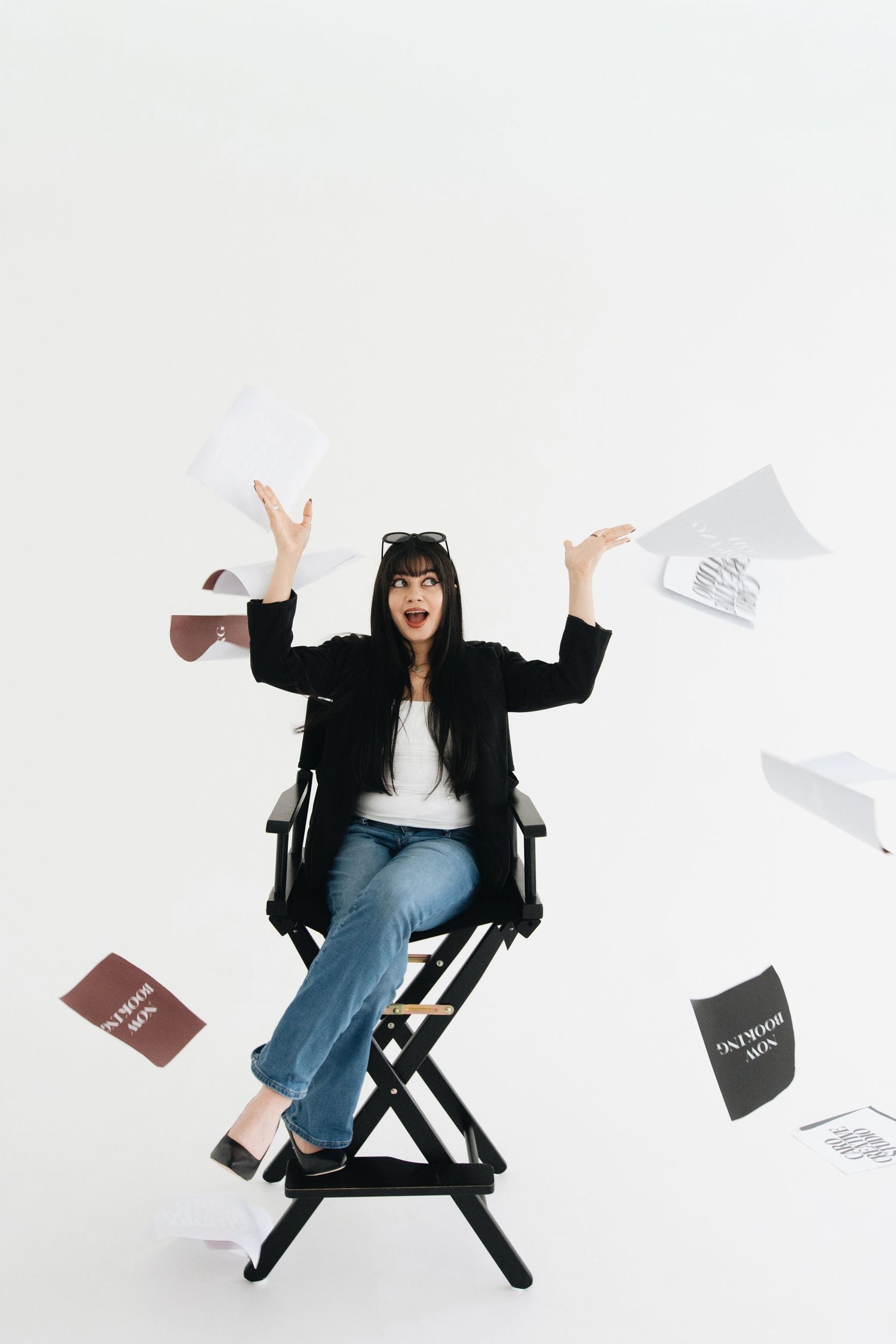 Excited woman in black blazer sits in director chair tossing papers in air against white background.