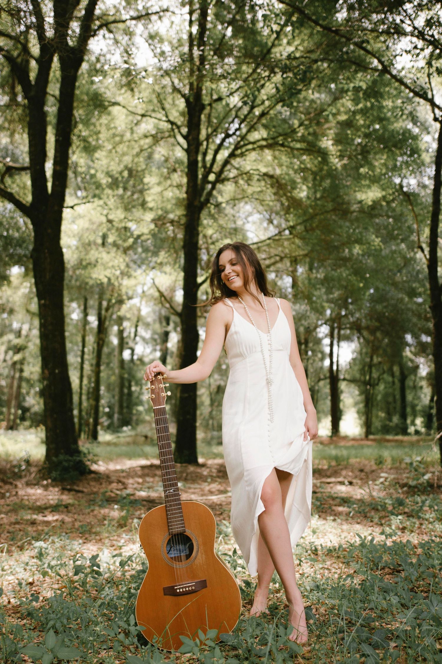 Artistic portrait session in a wooded area with acoustic guitar and flowing white dress.