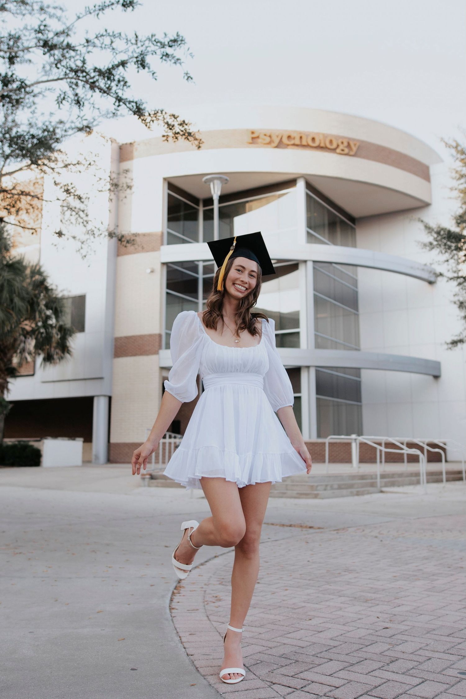 Graduate in white summer dress posing playfully outside a modern building.