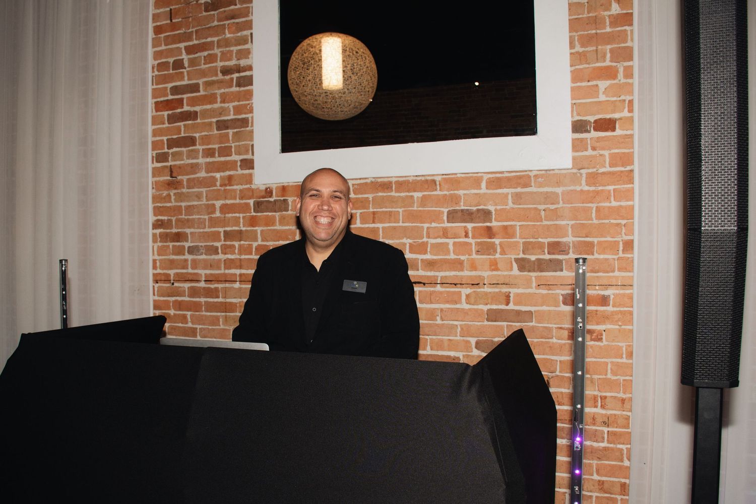 A DJ stands behind black equipment setup against an exposed brick wall with mood lighting.