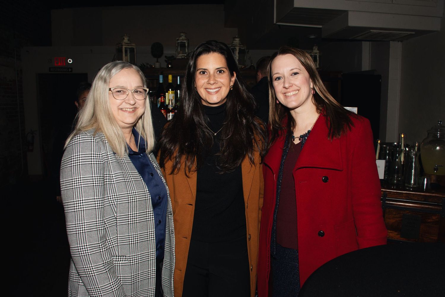 Three people in formal winter coats pose together at a dimly lit evening event.