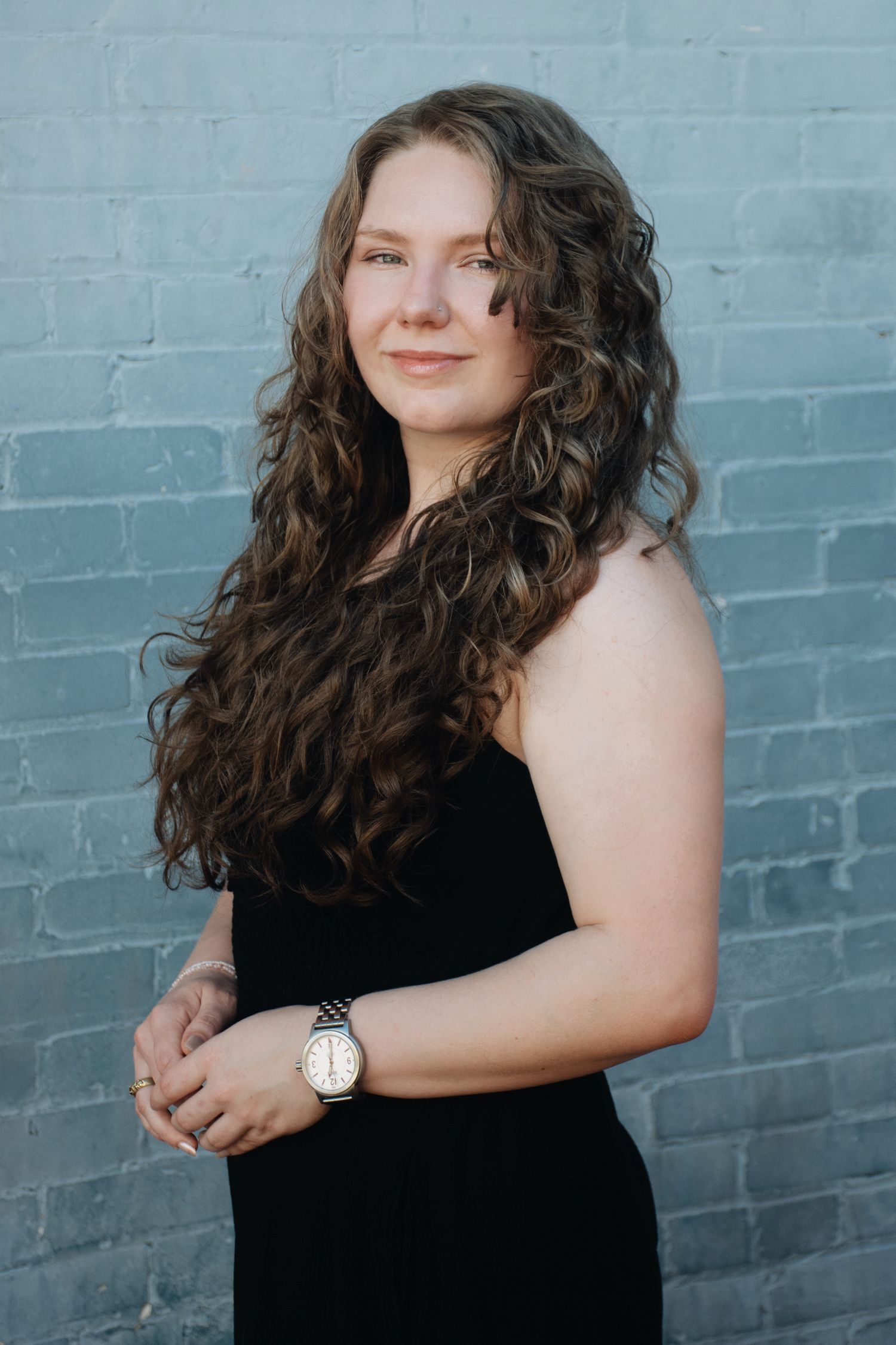 A woman in a black sleeveless top poses against a blue painted brick wall in a portrait series.