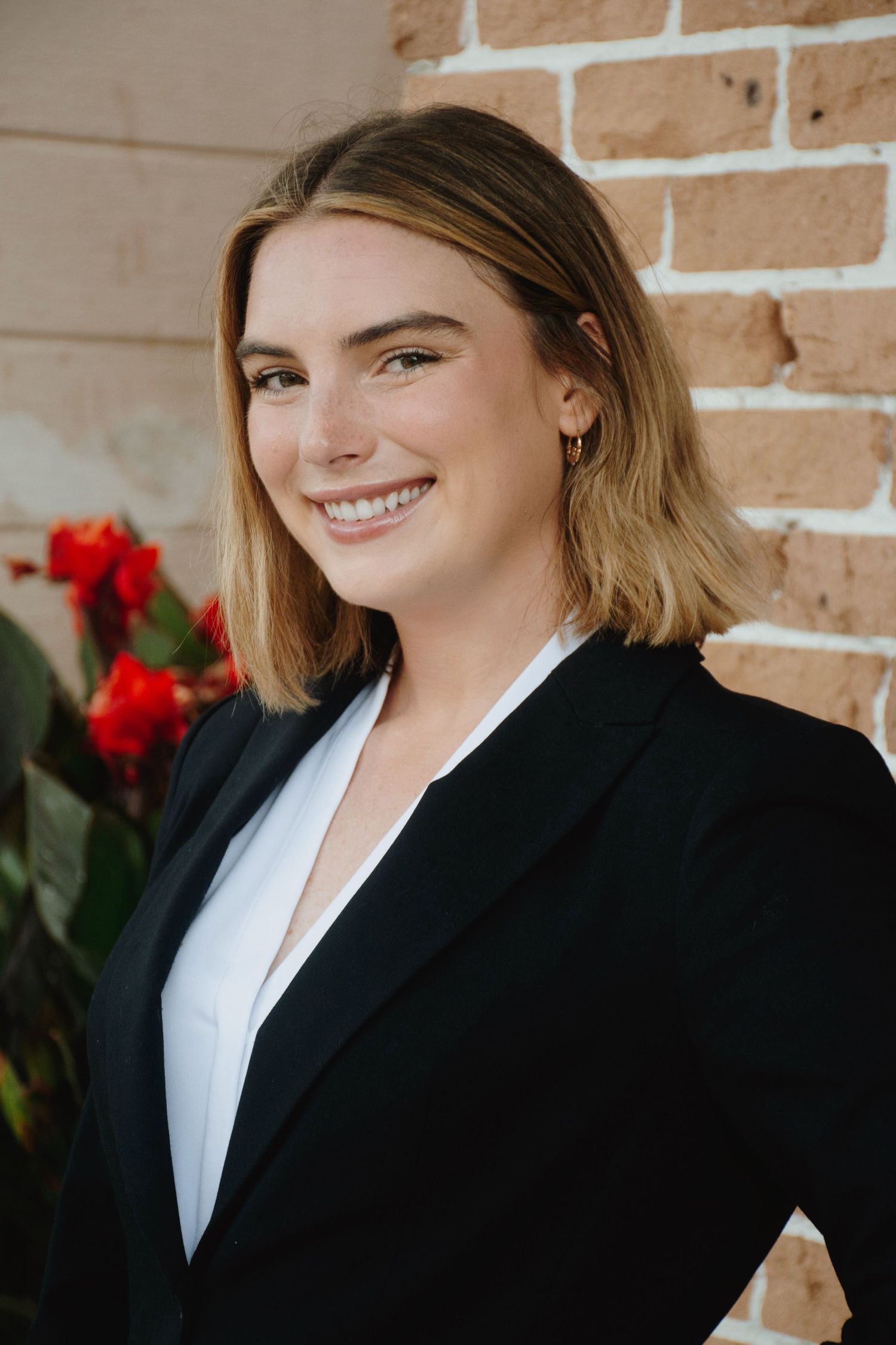 Professional headshot wearing a black blazer and white blouse against a brick wall.