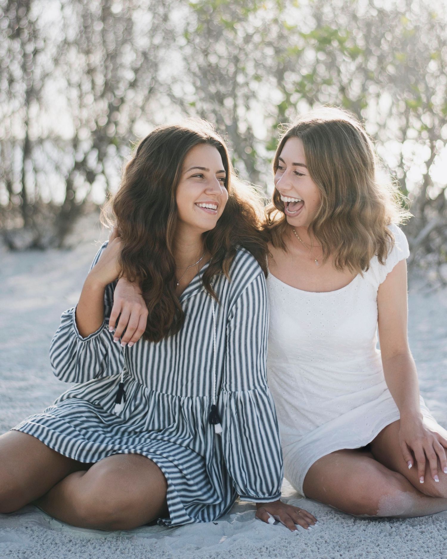 Two friends laughing together while lying on a sandy beach in casual summer outfits.