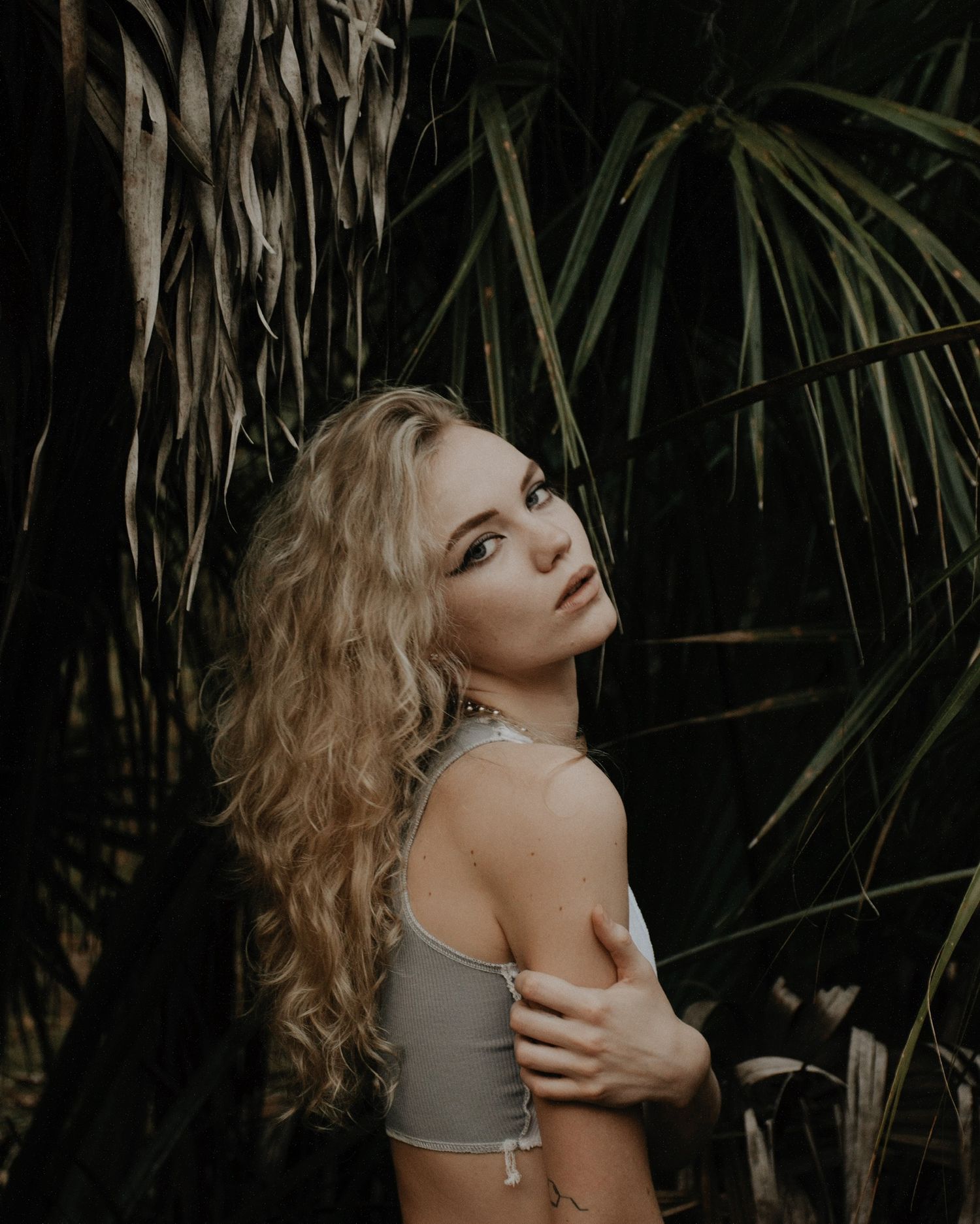 Moody fashion photoshoot with model in gray top against dark palm leaves.