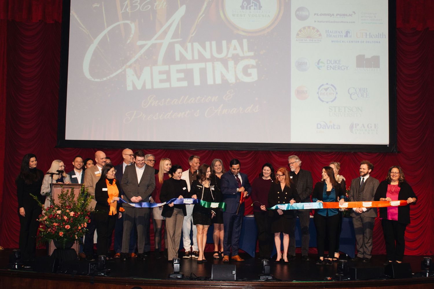 Business conference attendees gather on stage during an awards ceremony with red curtain backdrop and projection screens.