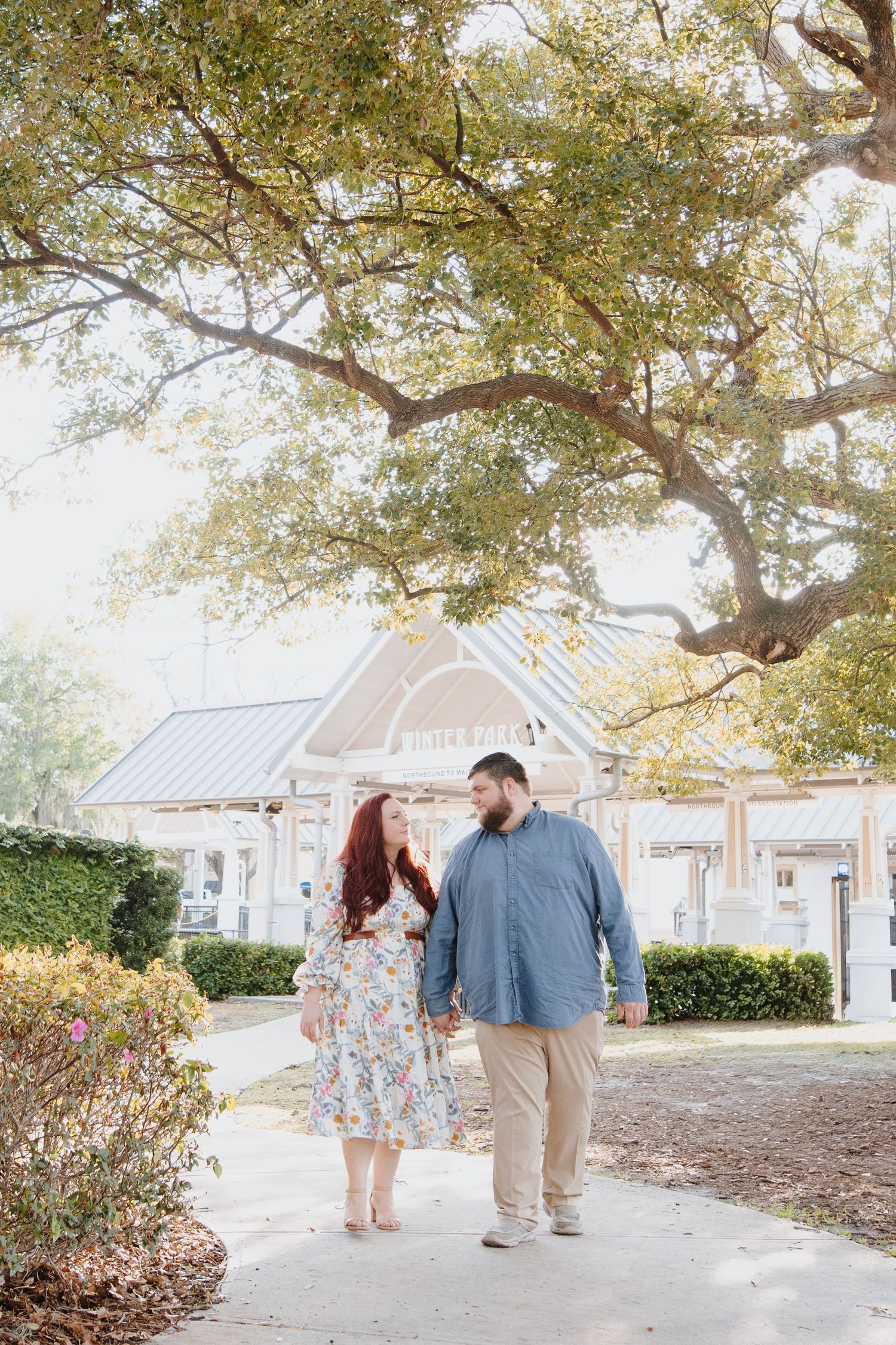 A couple shares romantic moments together on a walkway under sunlit trees near white buildings.