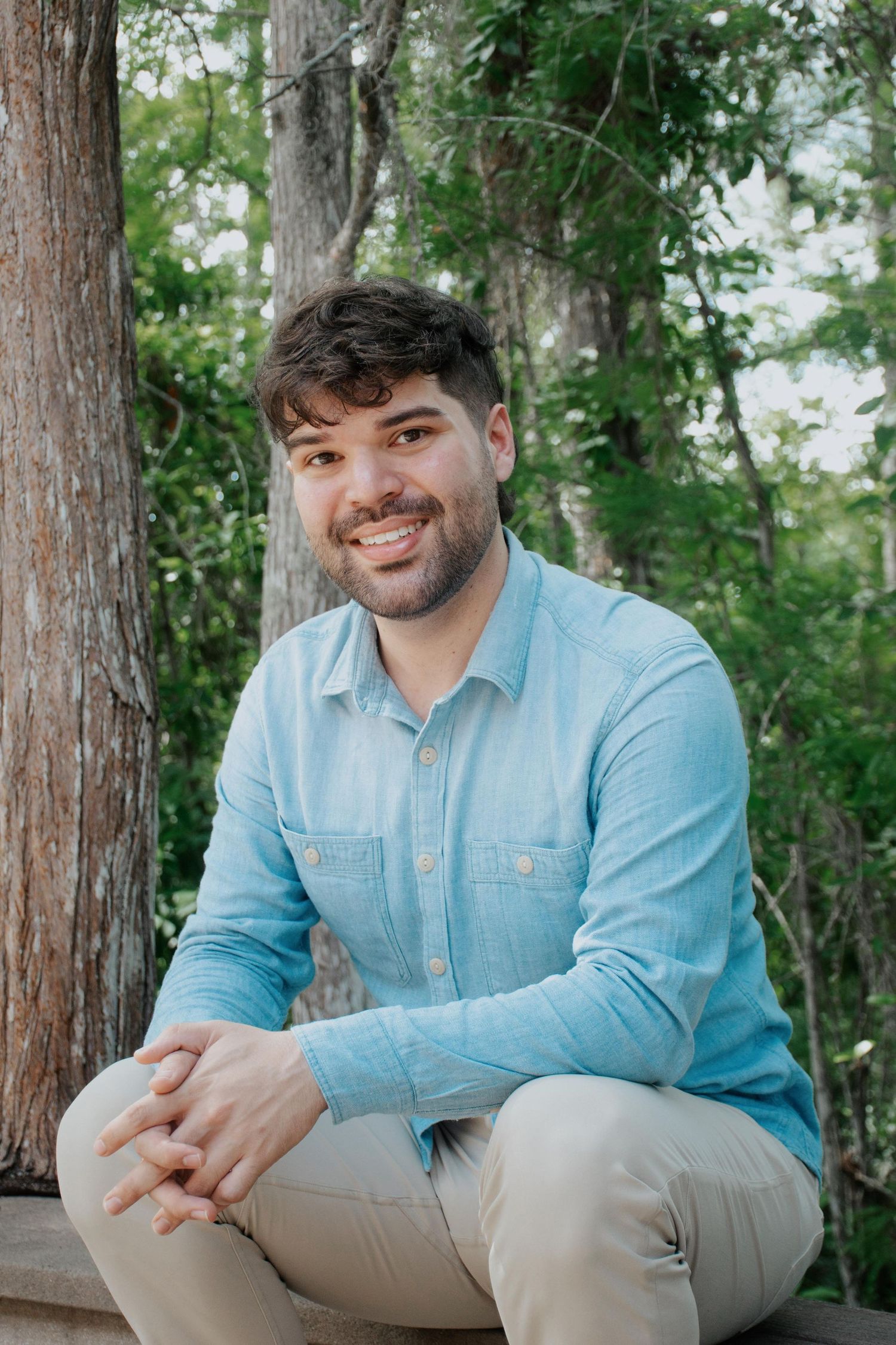 Graduate in light blue shirt sits casually on wooden bench surrounded by trees.