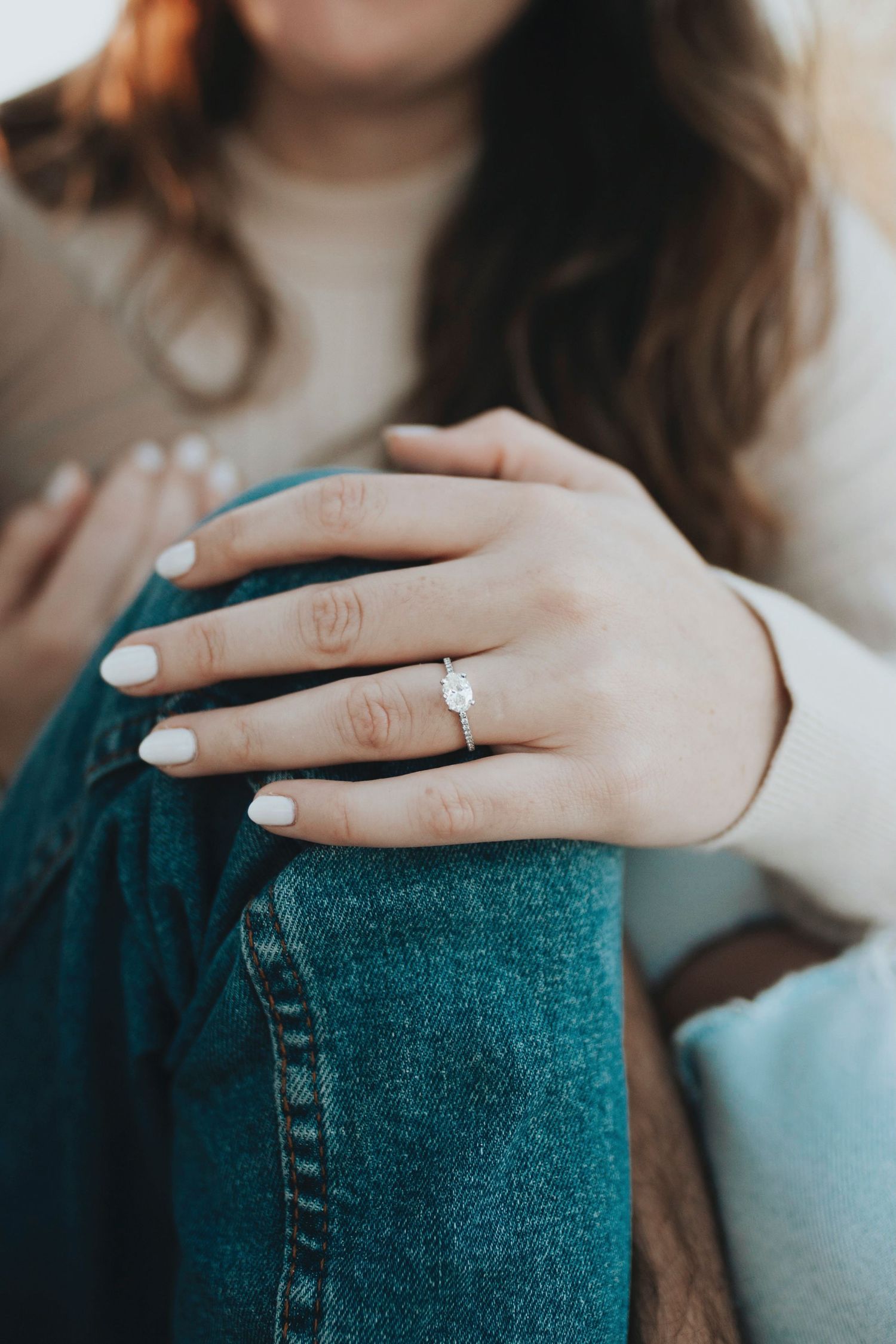 Close-up of a hand wearing a white pearl engagement ring with light blue nail polish against teal denim.