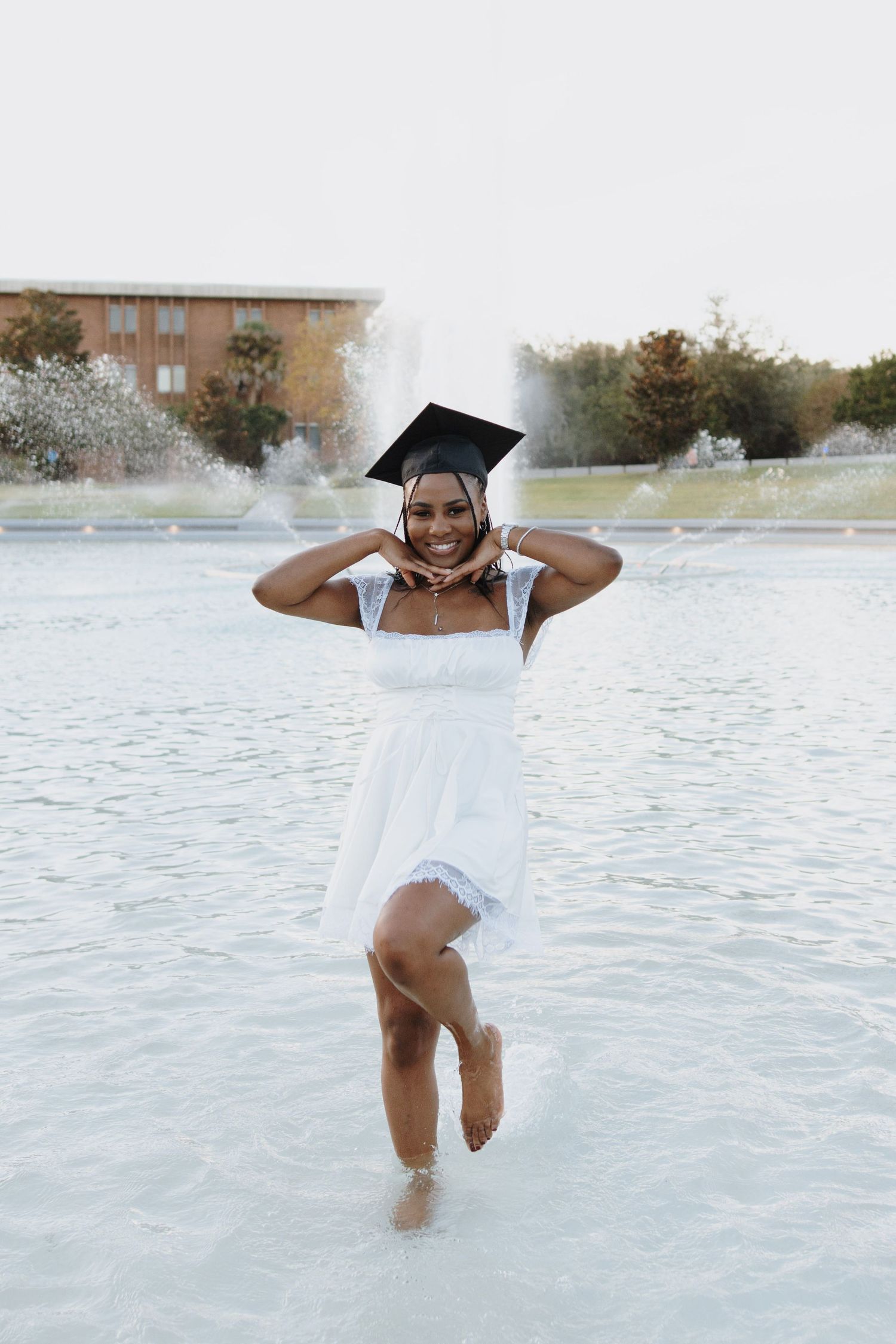 A graduate in white dress splashes in fountain water while wearing graduation cap.