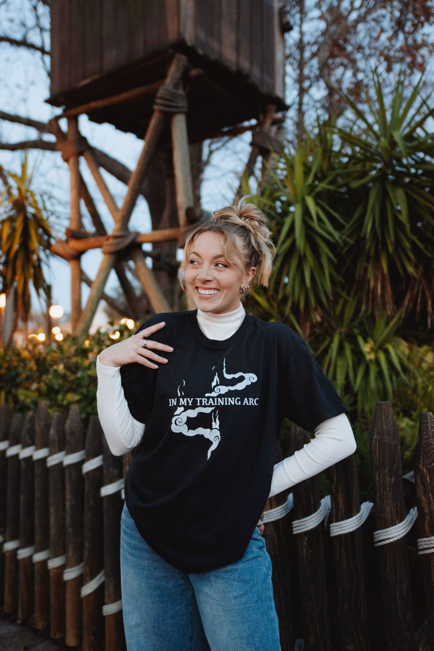 Person in black sweatshirt and jeans poses expressively against wooden fence at sunset.