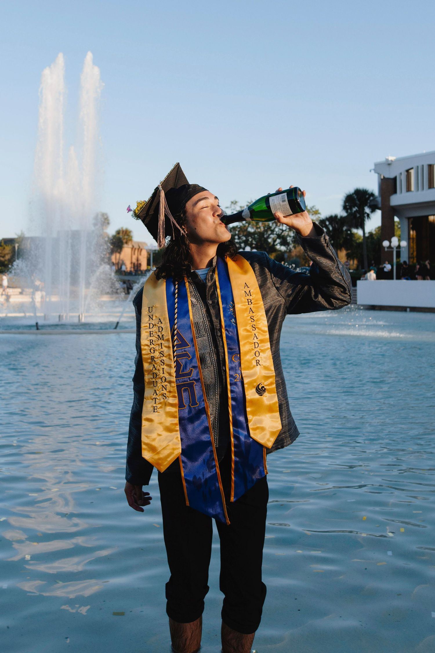 Graduate in regalia drinks from bottle in front of fountain during celebration.