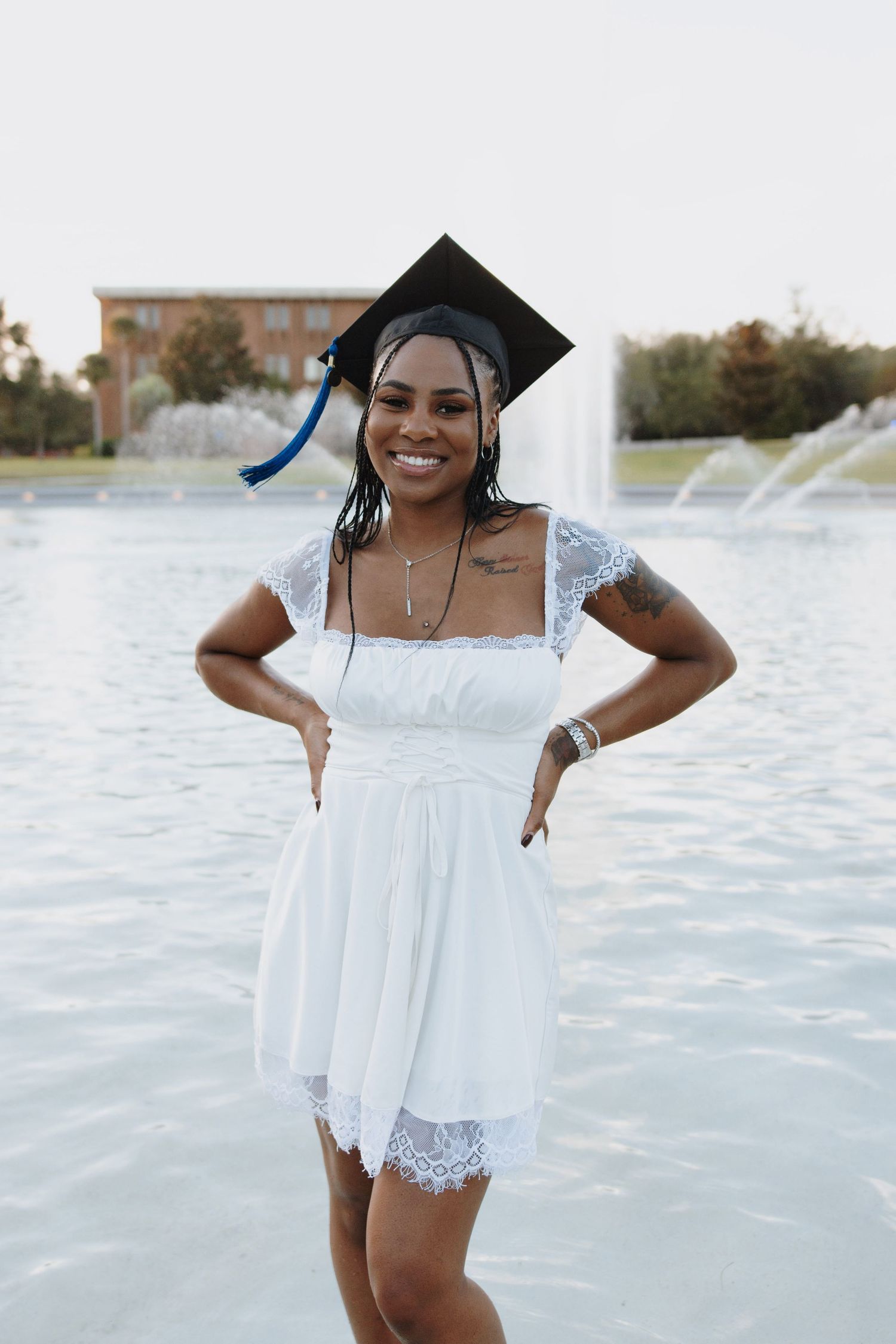 A graduate in white lace dress poses joyfully by a fountain during graduation photoshoot.