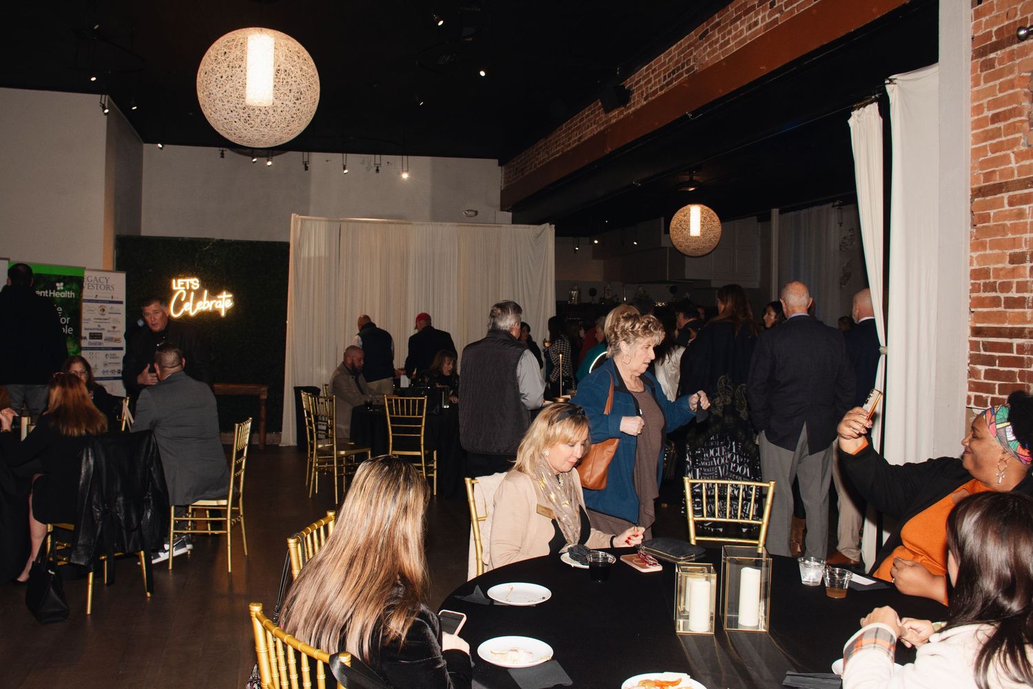 People gather in a dimly lit event space with round tables and hanging pendant lights creating an intimate atmosphere.