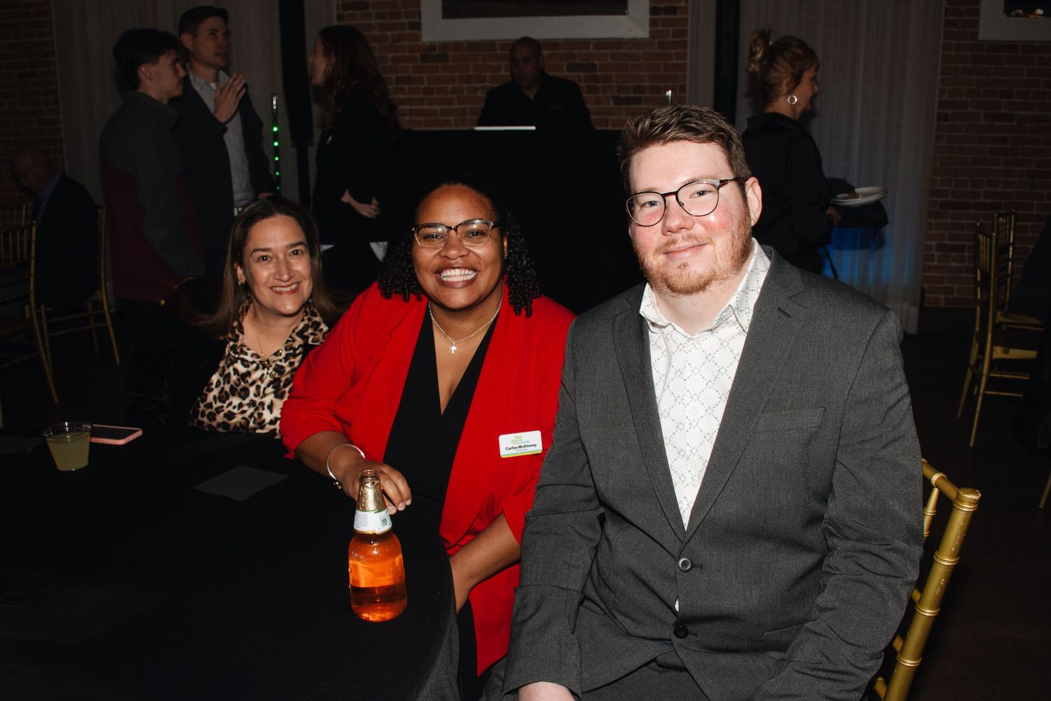 A group of people in formal attire gathered at an indoor event space with brick walls and dark lighting.