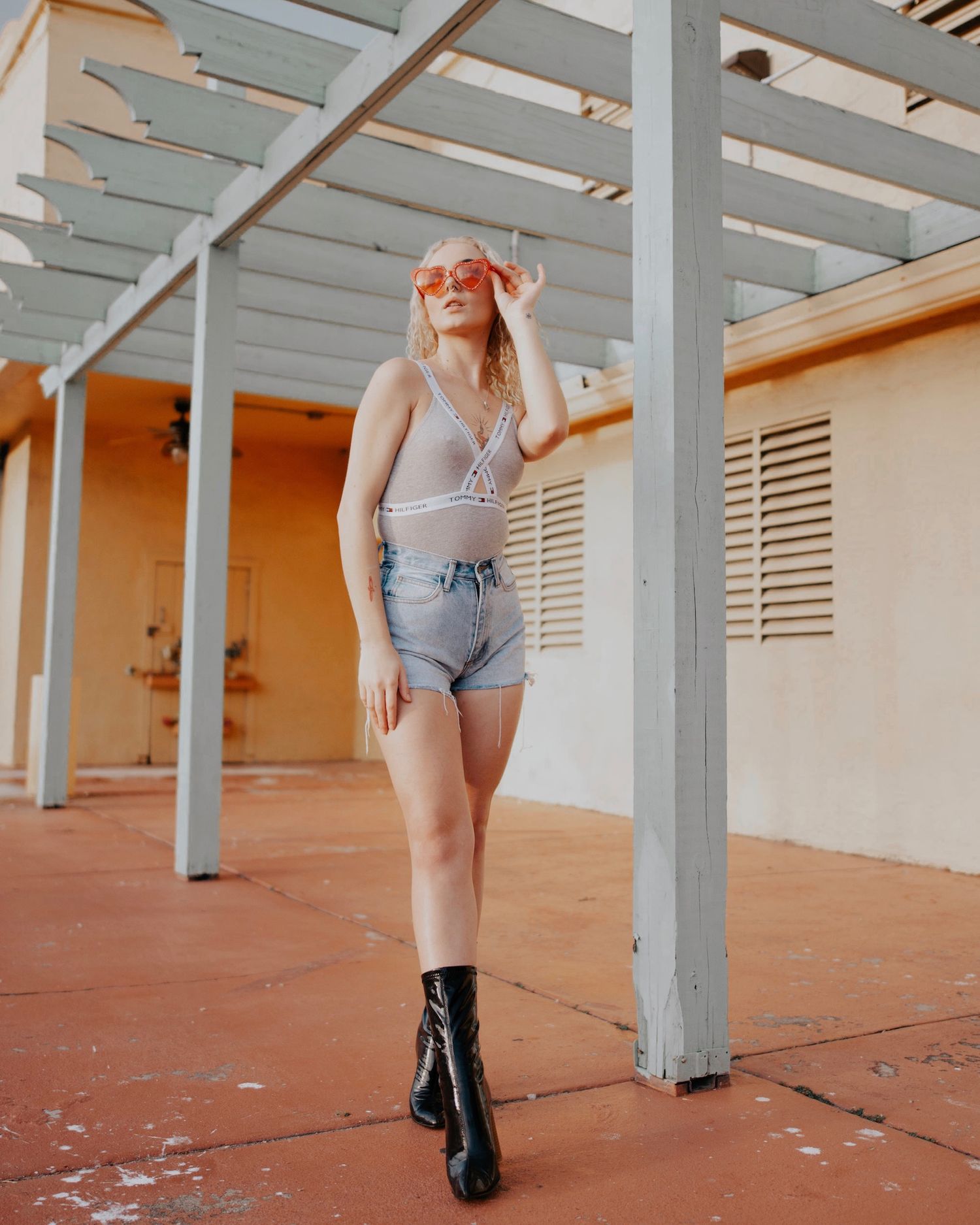 Model in denim shorts and boots poses under industrial ceiling beams and orange walls.