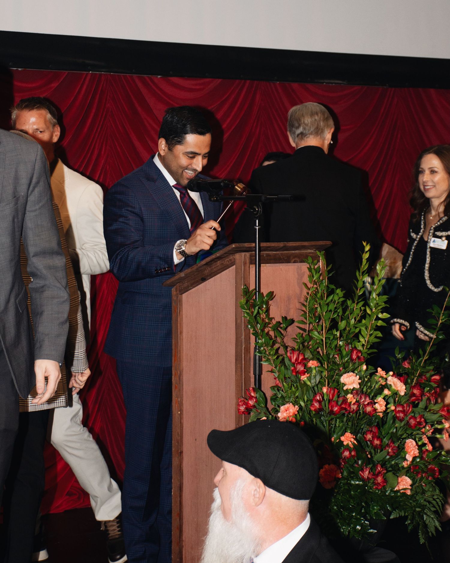People gathered at an event with red curtains and floral arrangements in the background.