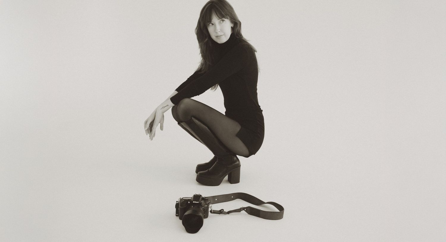Woman in black turtleneck crouching beside a camera on white background in a stylish monochrome portrait.