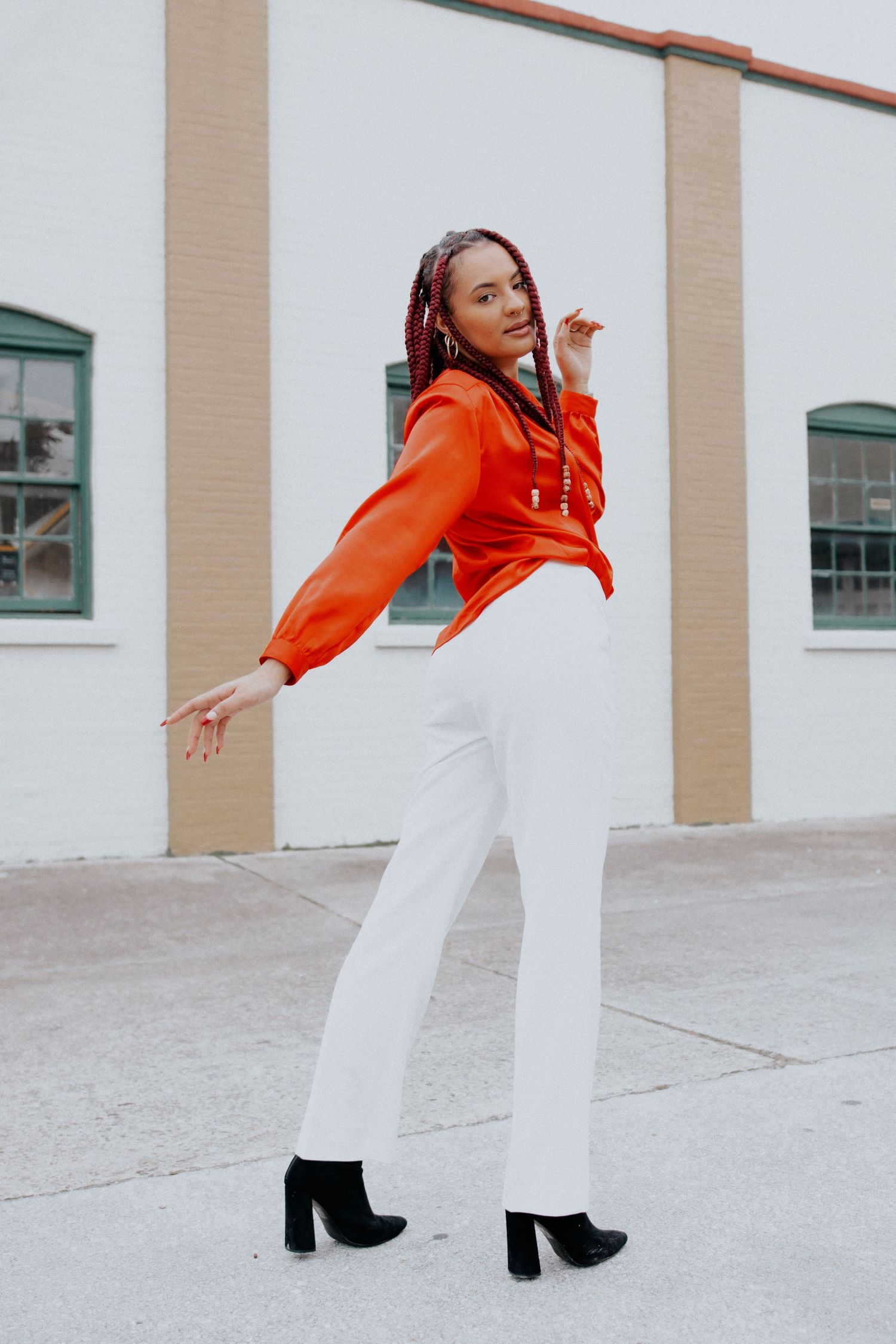 Fashion model wearing bright orange top and white pants poses dynamically against neutral building exterior.