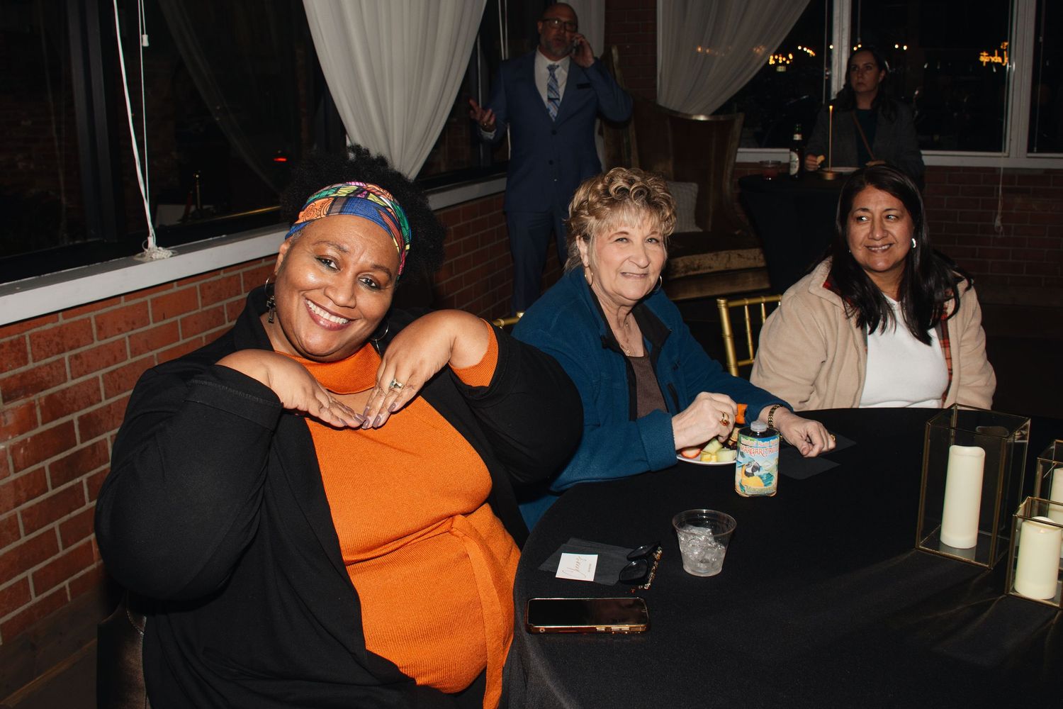 Three people sit together at a table during an evening event with brick walls and curtains visible in the background.