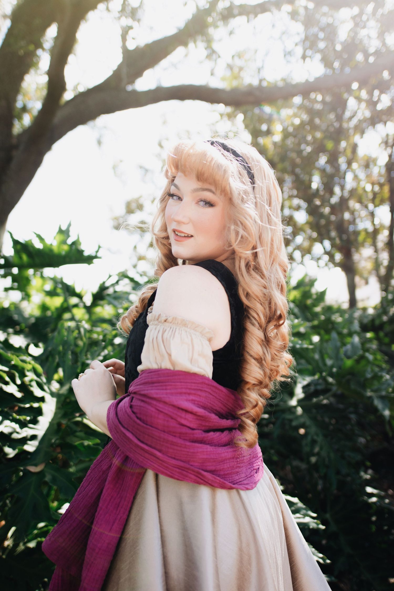 Person in vintage dress with burgundy scarf poses playfully against garden backdrop.