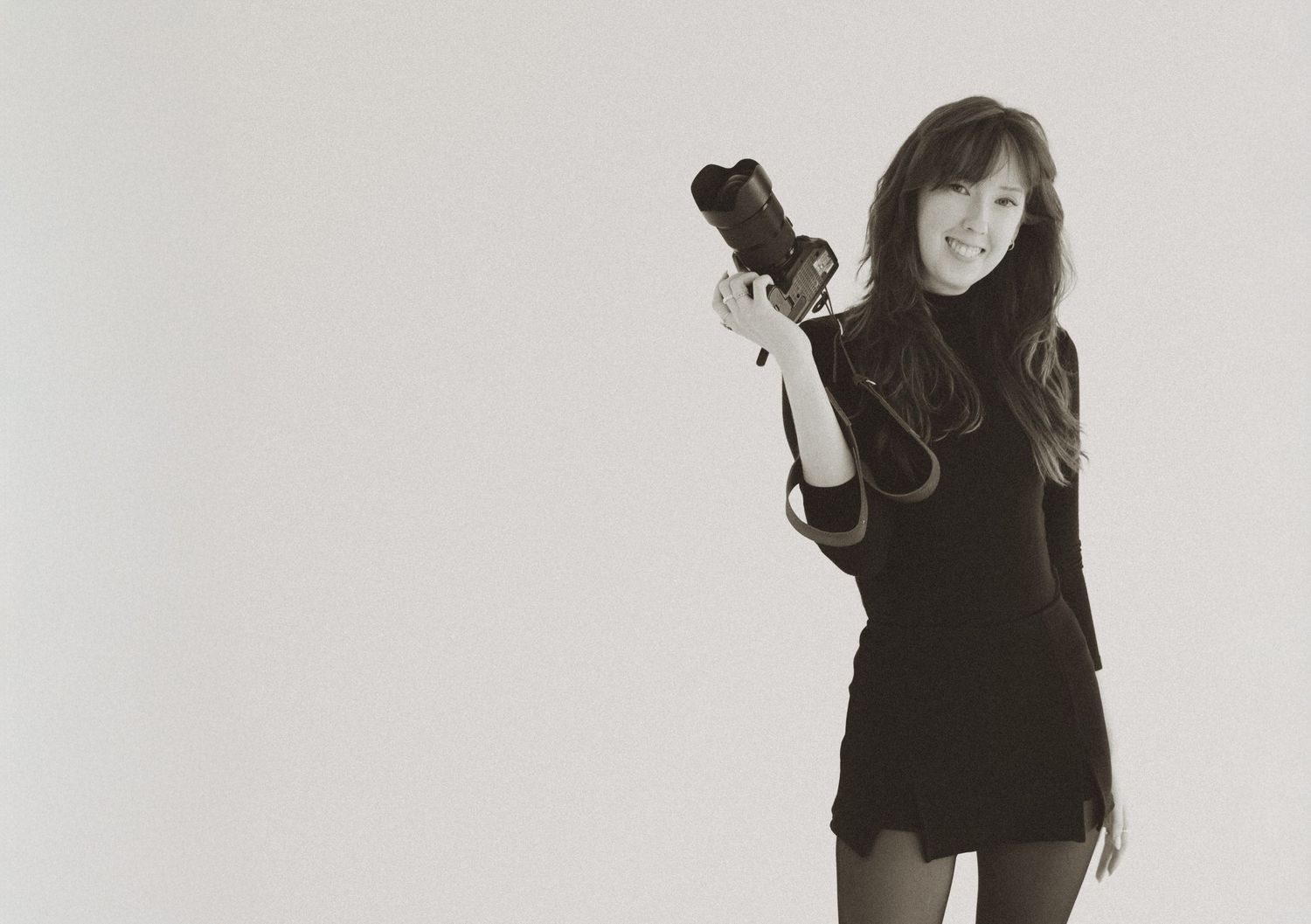 Young woman in black outfit holding a camera, posing in a black and white studio photo.