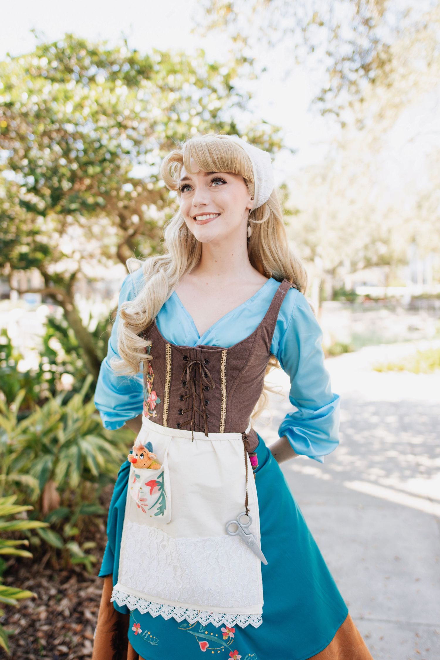 A costumed character in a turquoise dress and brown corset poses in a sunny garden setting.