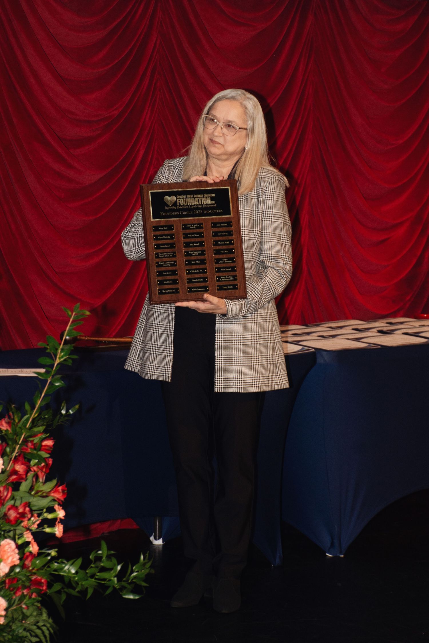Award ceremony presentation with wooden plaques against red curtain backdrop and decorative floral arrangements.