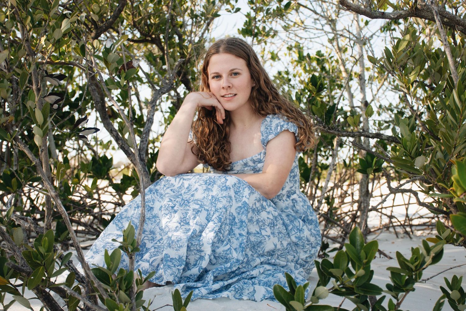 Young woman in blue floral dress poses among leafy trees in outdoor portrait session.