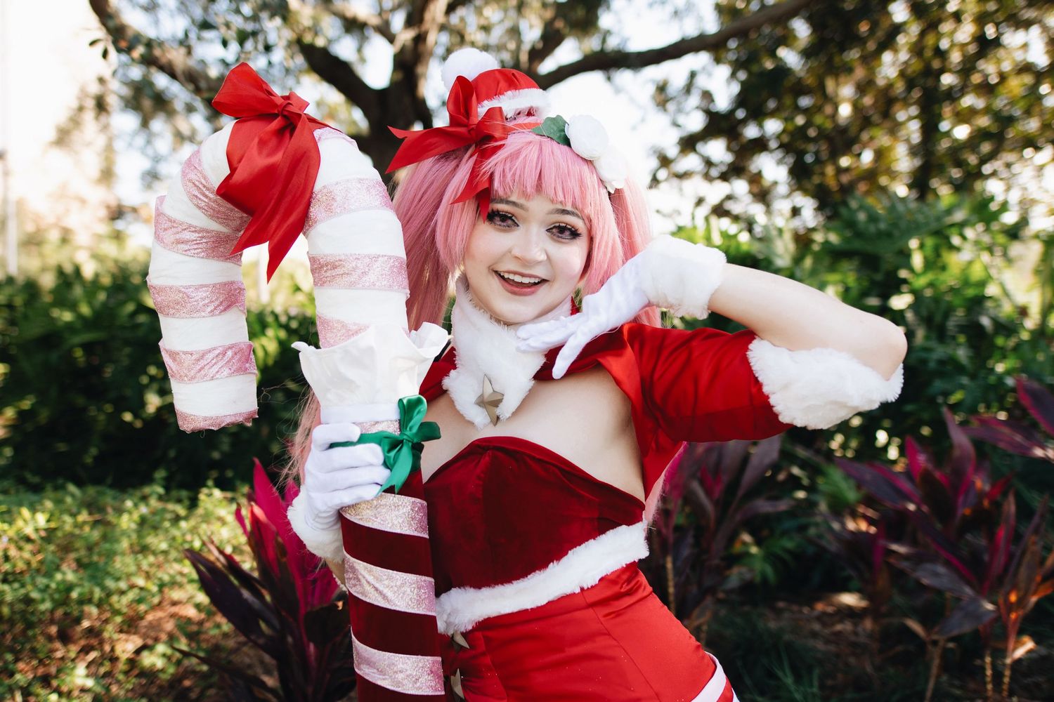 Cosplayer in a festive red and white Christmas-themed dress poses playfully in an outdoor garden setting.