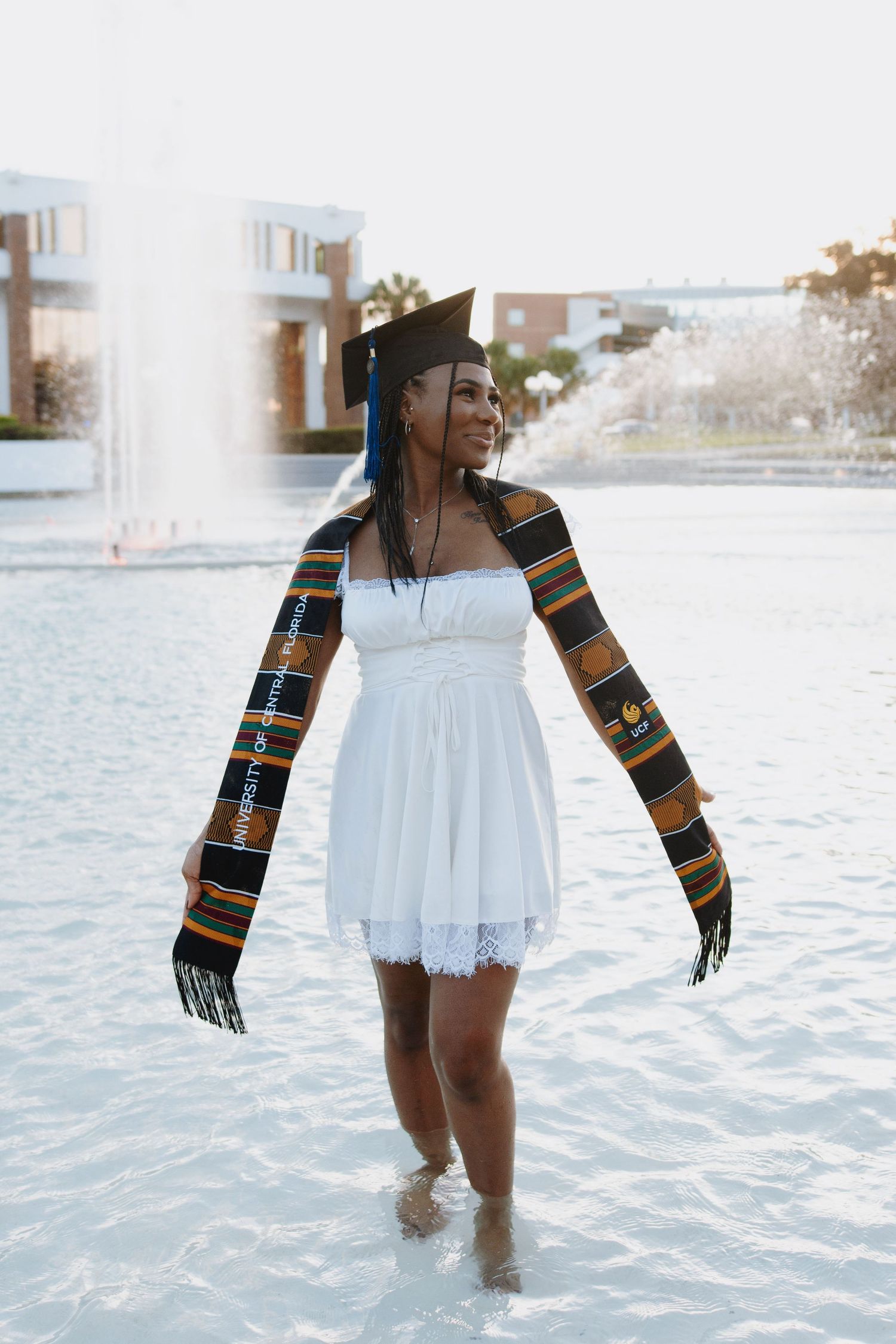 A graduate in white dress and patterned stole wades through fountain water during celebration photos.