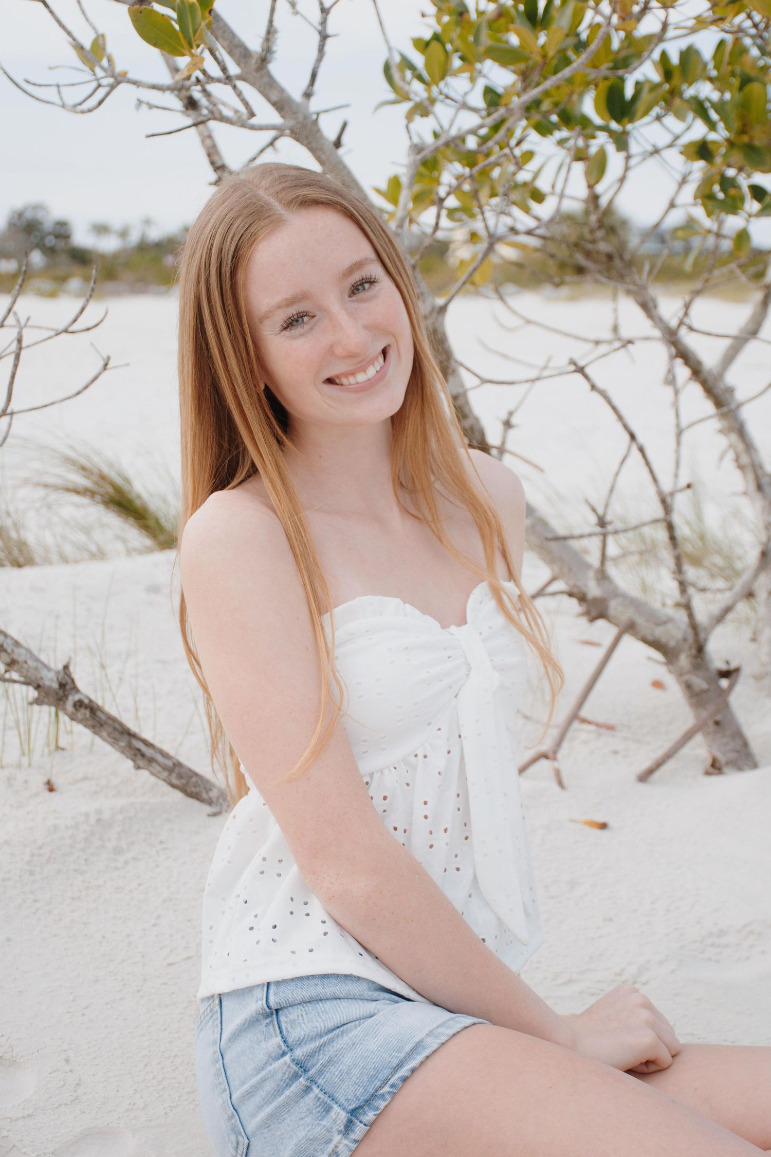 Young woman in white top poses on beach with tree branches in background.