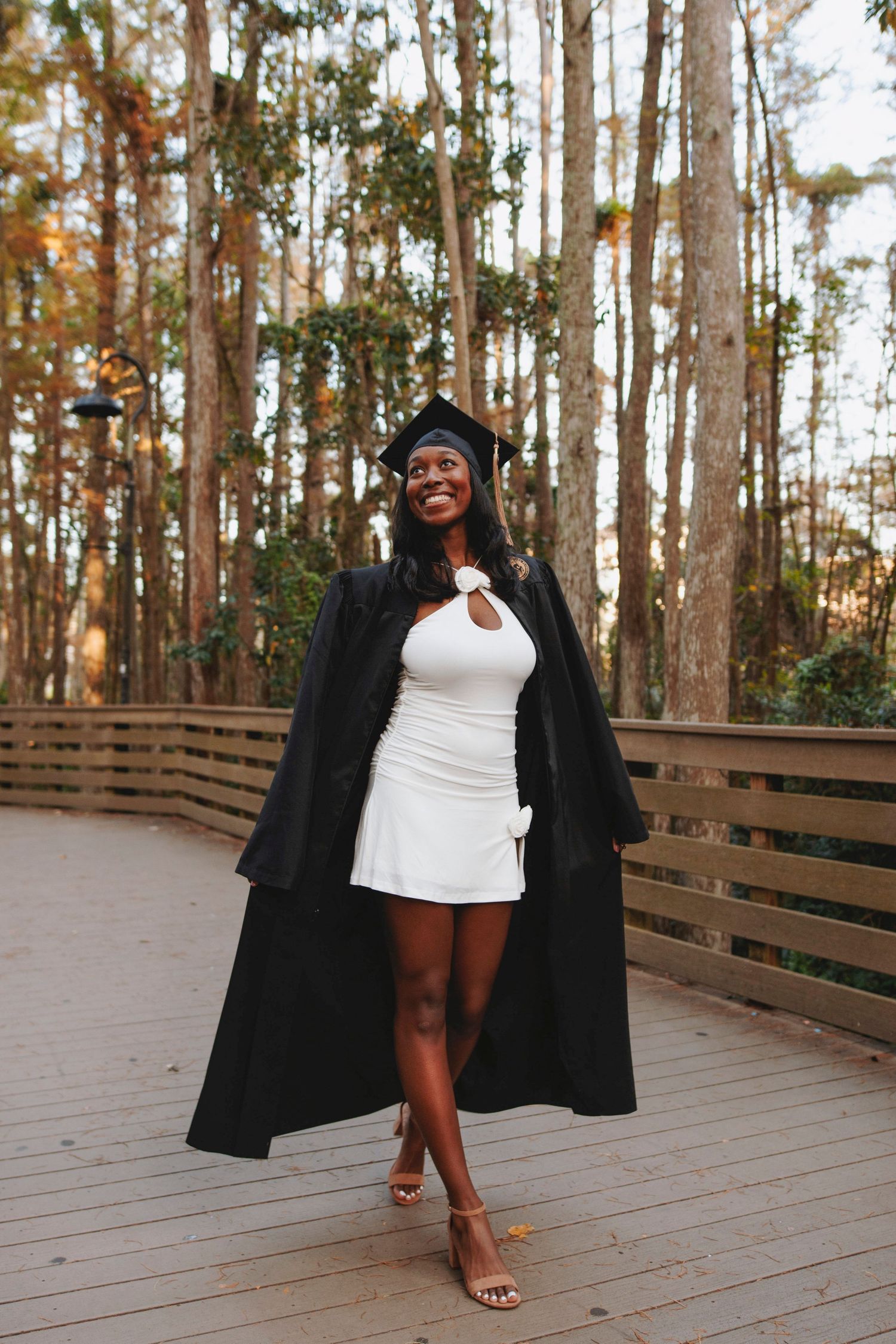 Graduate in black cap and gown poses on wooden deck surrounded by tall pine trees.