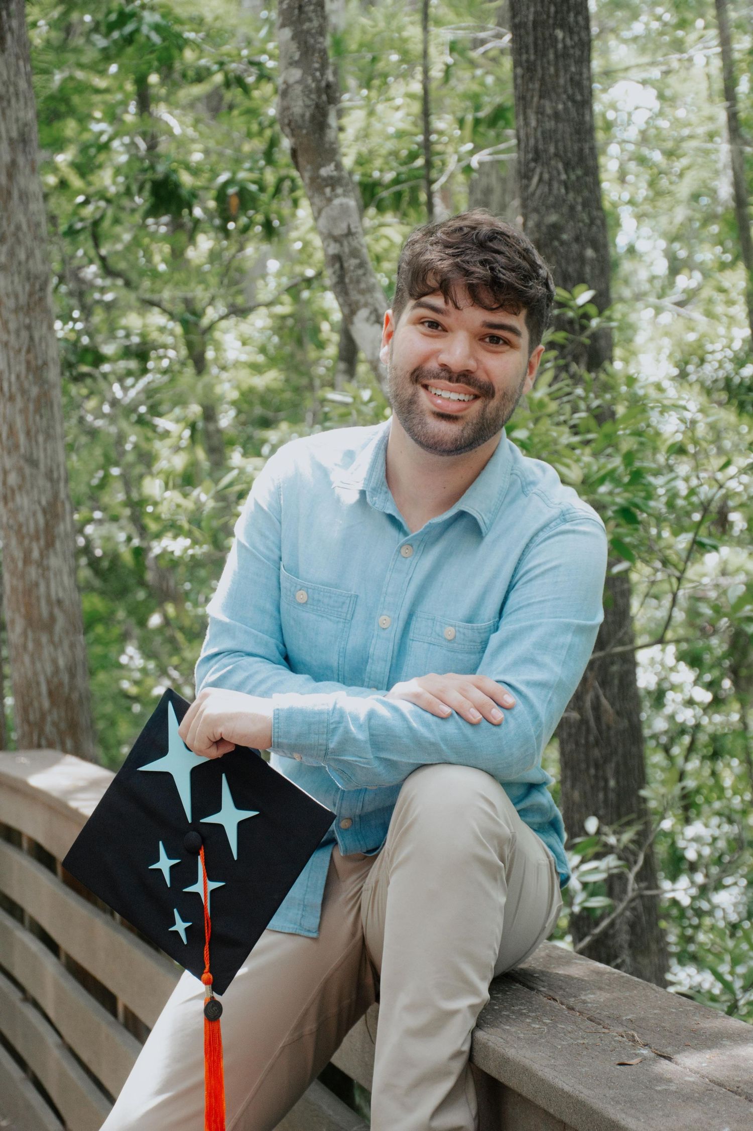 A person in a light blue button-down shirt sits casually on a wooden bench in a wooded outdoor setting.