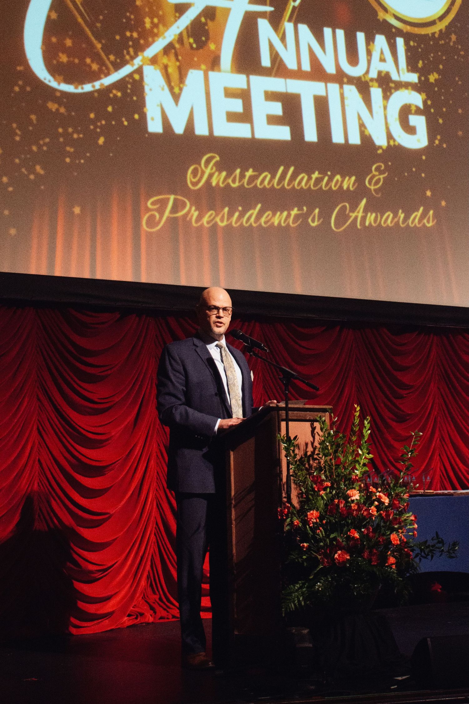 A speaker stands at a podium on stage with red curtains and illuminated text display during an annual meeting.