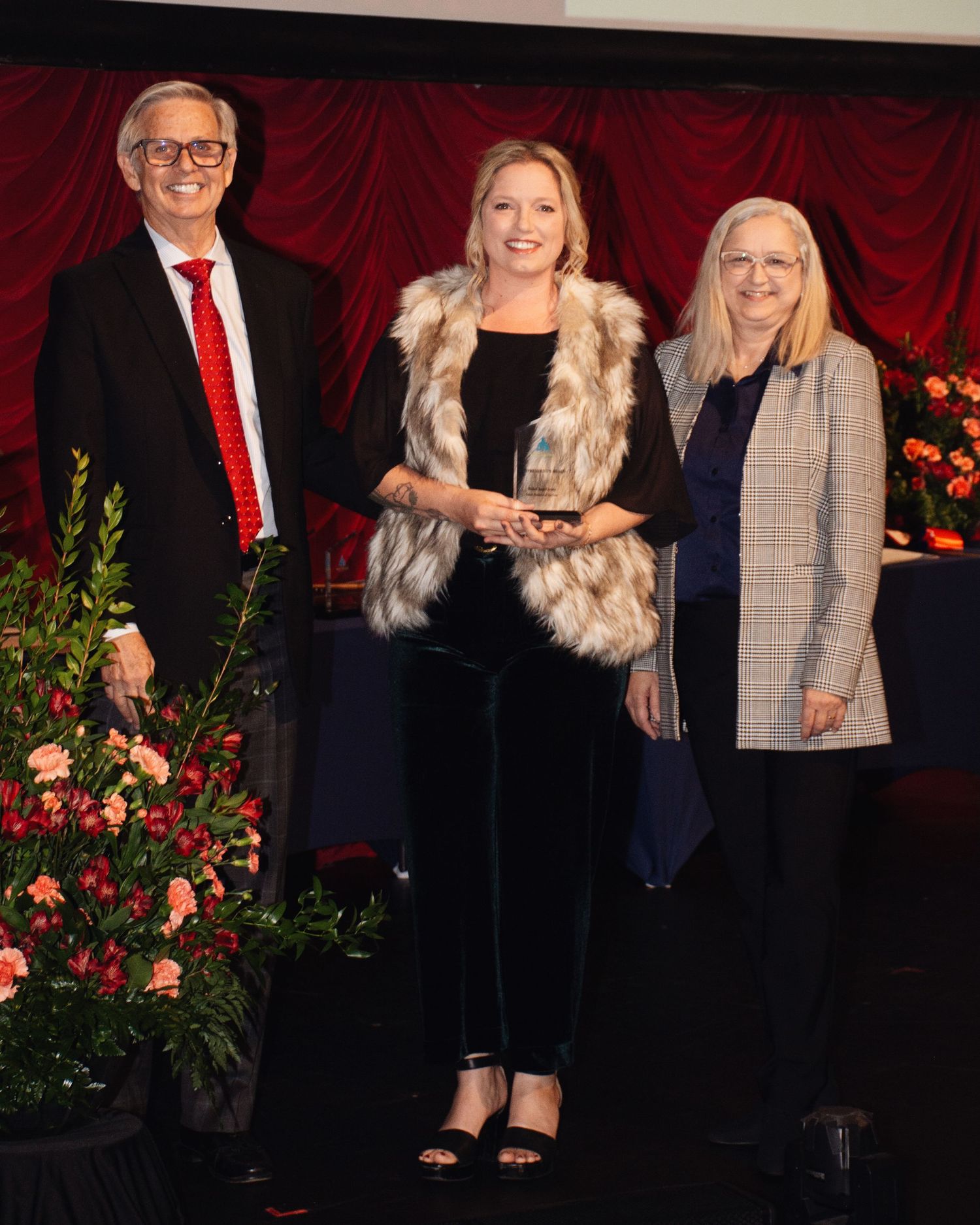 Three people in formal attire stand together at an awards ceremony with red curtain backdrop and floral decorations.