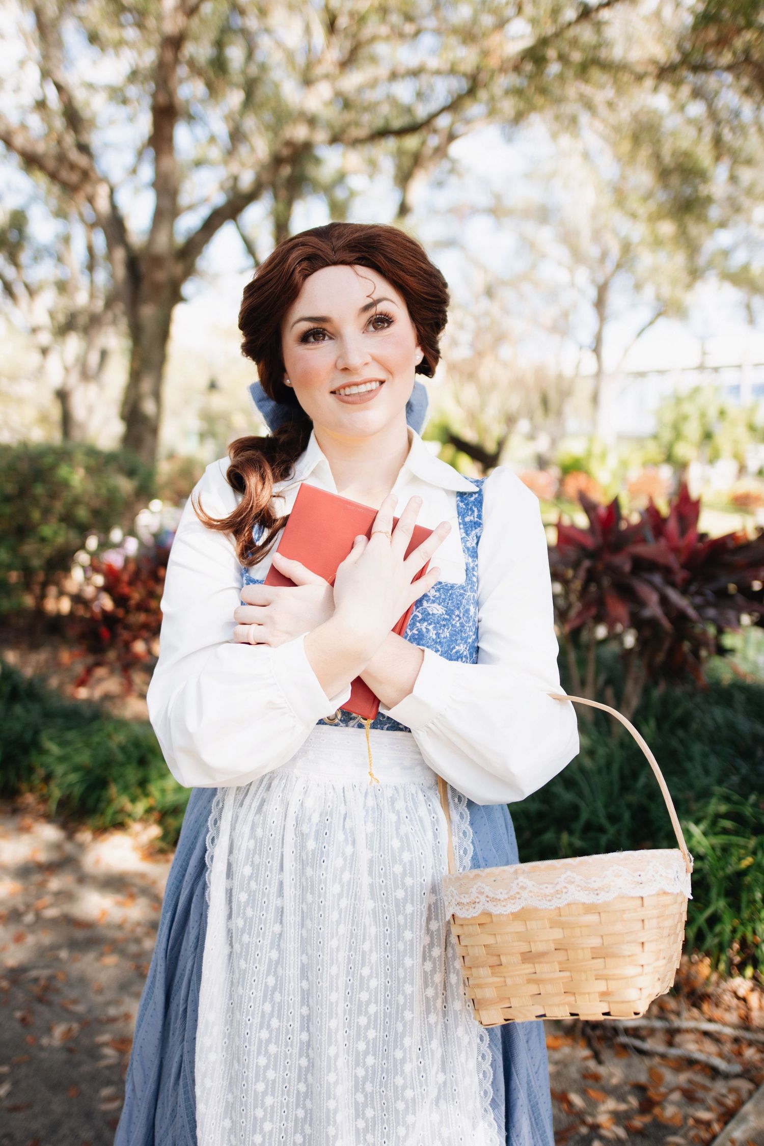A figure in a blue provincial dress holds a red book and wicker basket in a garden setting.