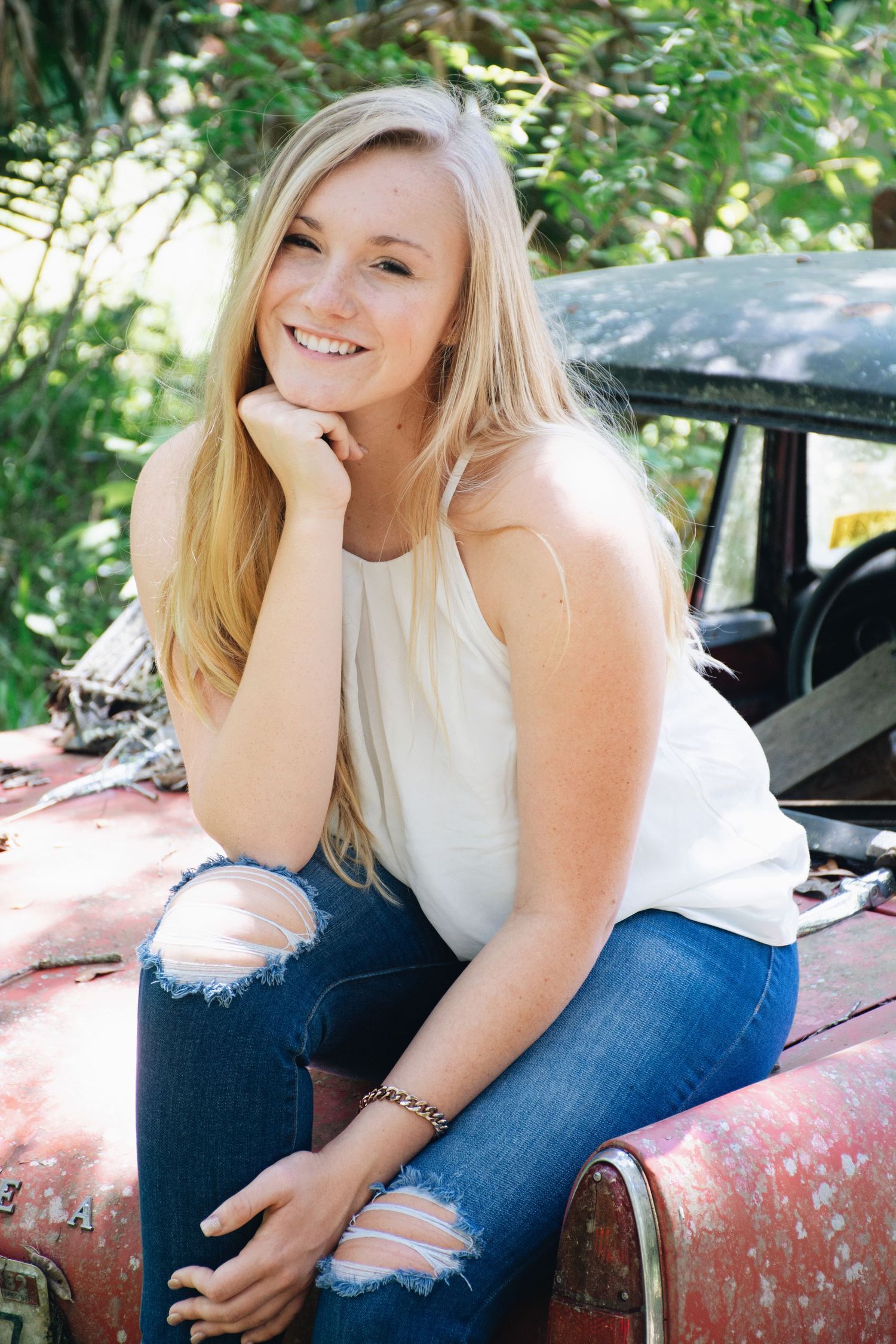 A person in a white top and ripped jeans poses casually on a vintage red car in an outdoor setting.