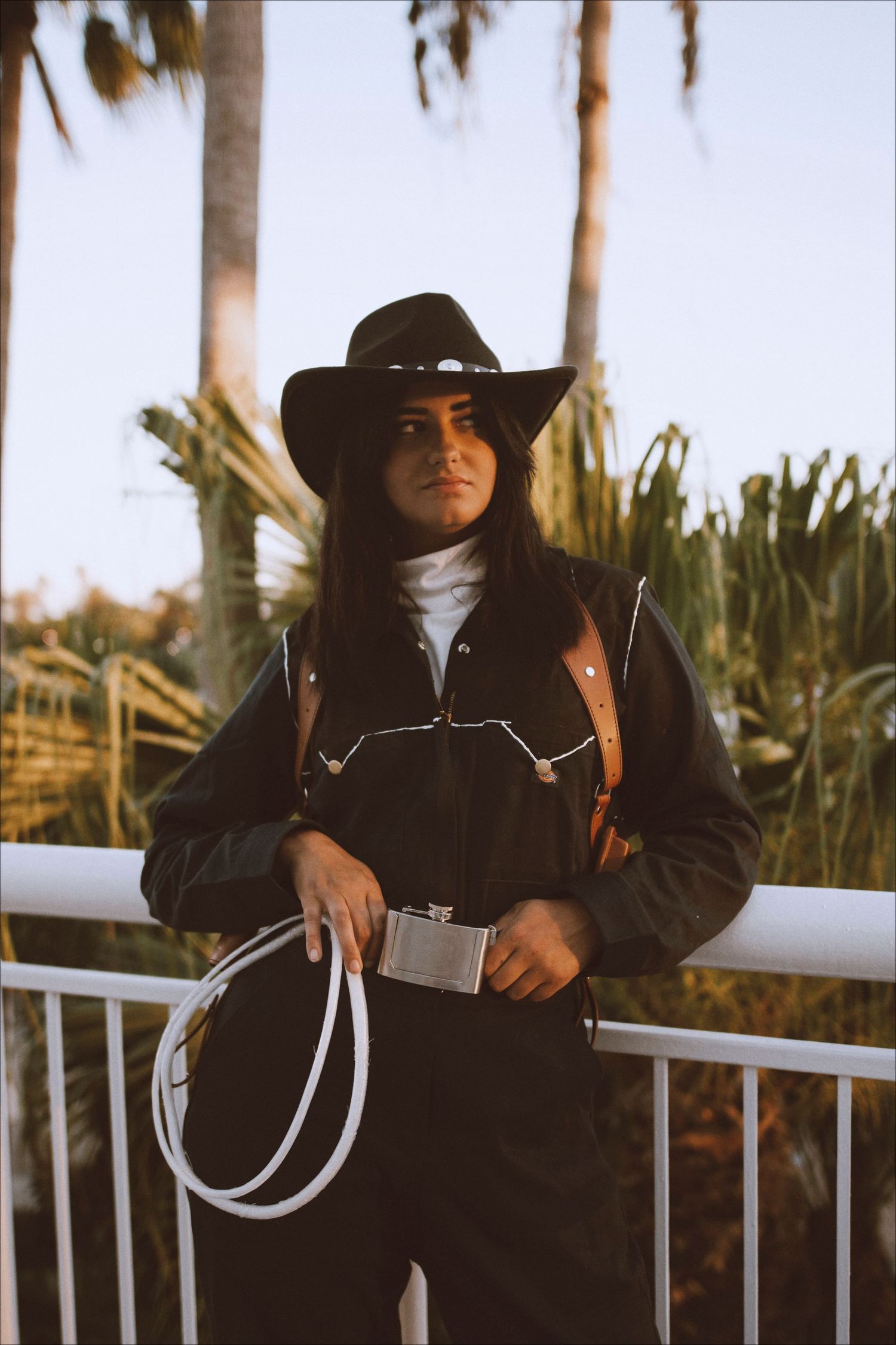 Western-inspired cosplay photoshoot featuring black outfit and cowboy hat against palm trees at sunset.