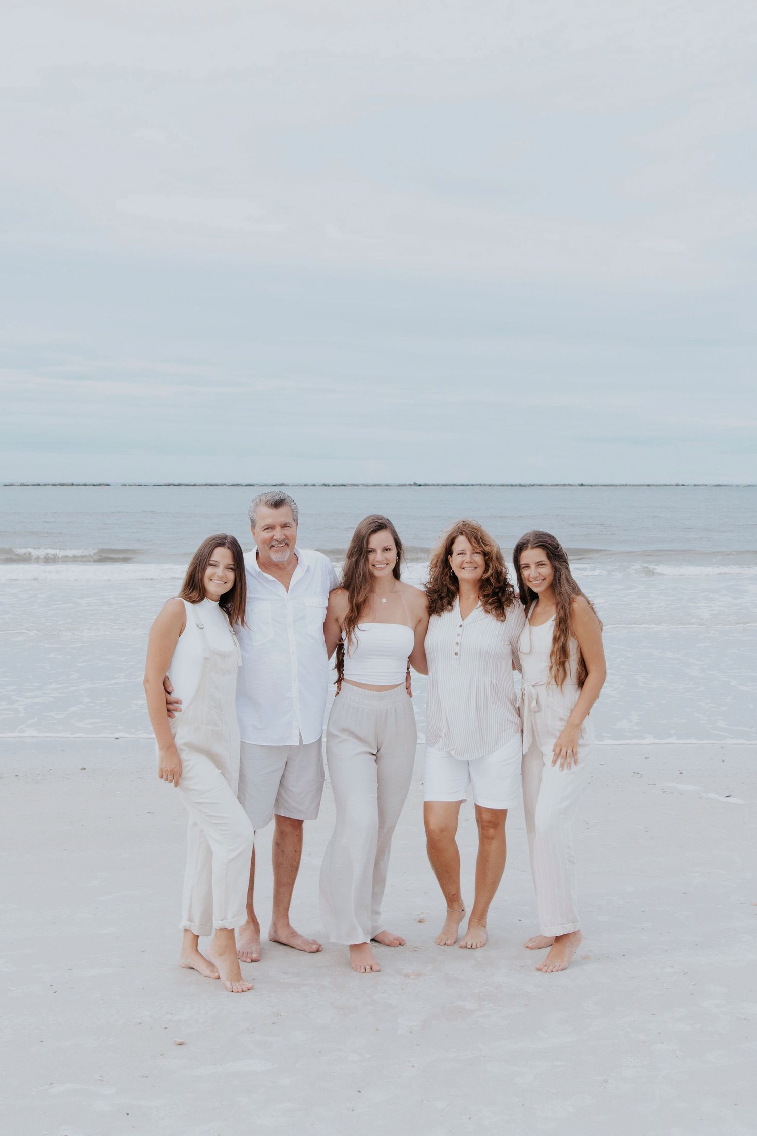 A group in coordinated white and beige outfits poses together on a pristine beach with ocean waves in the background.