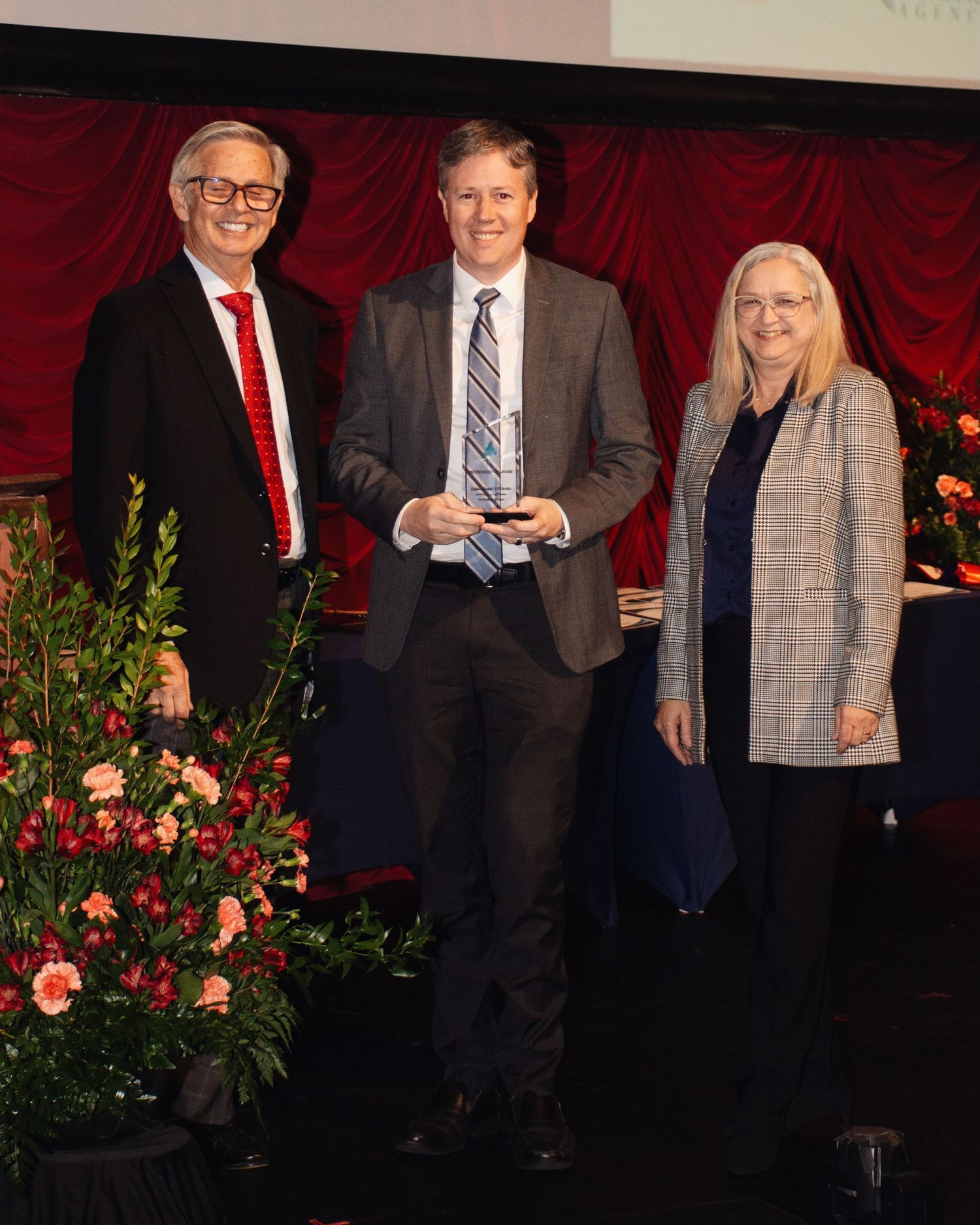 Three people in formal attire stand together on a stage with red curtains and floral decorations in the background.