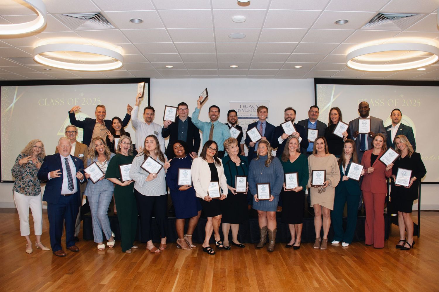 A large group of professionals holding awards and certificates pose together in a bright conference room with circular ceiling lights.