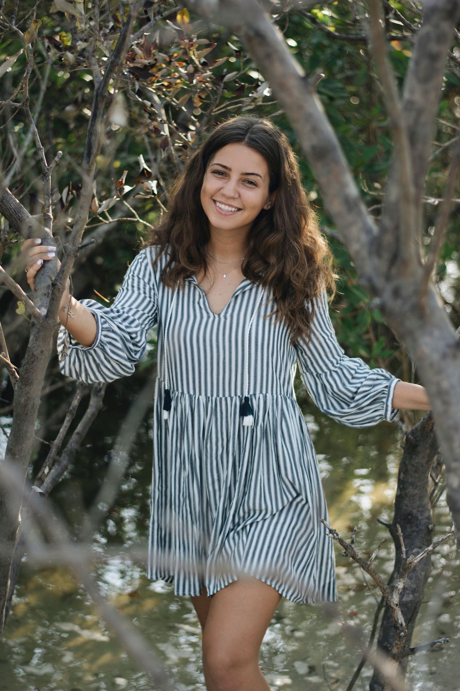 Person in striped dress posing playfully between trees in a natural outdoor setting.