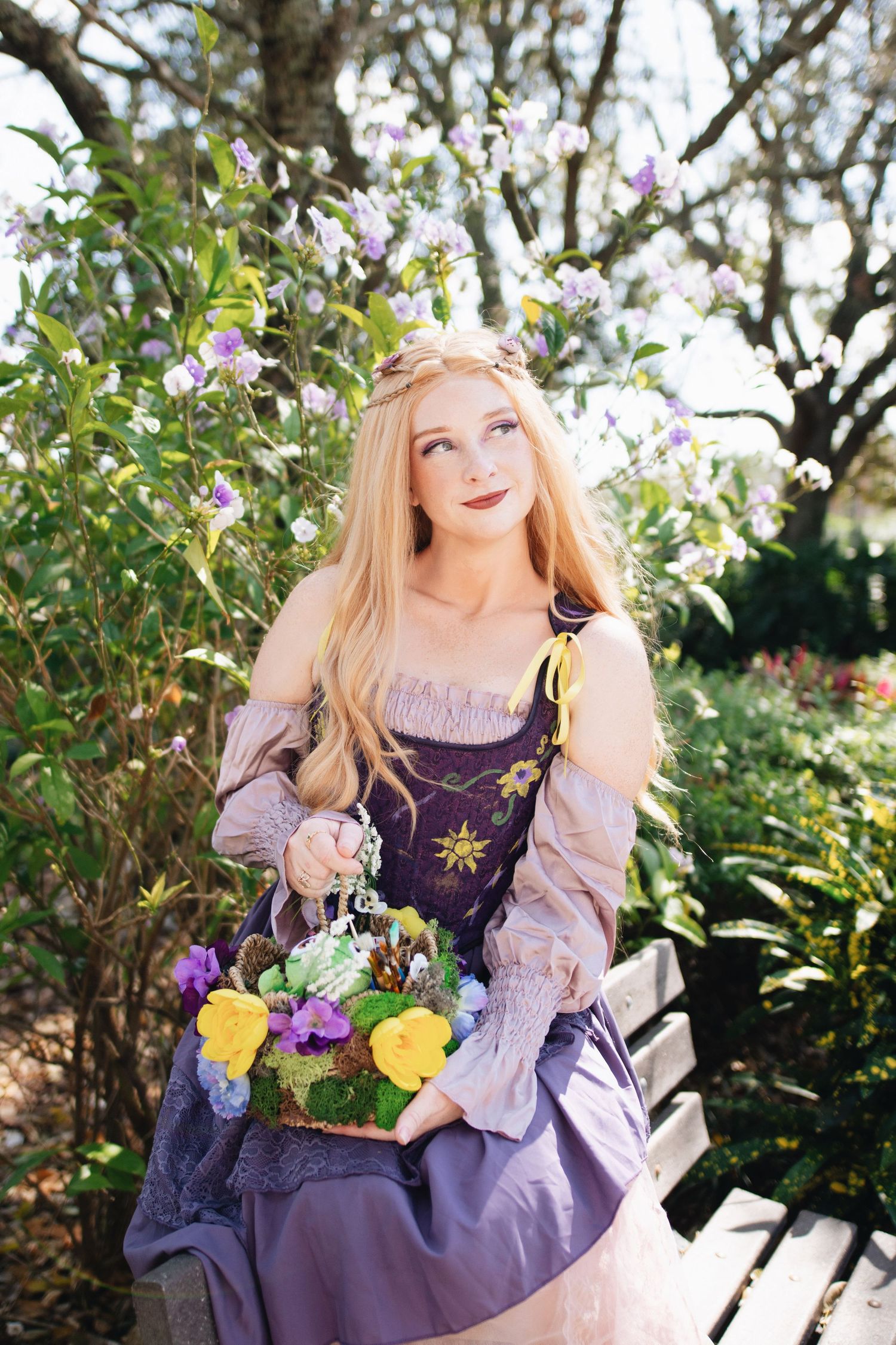 A cosplayer in a purple renaissance dress poses with flowers on a garden bench.