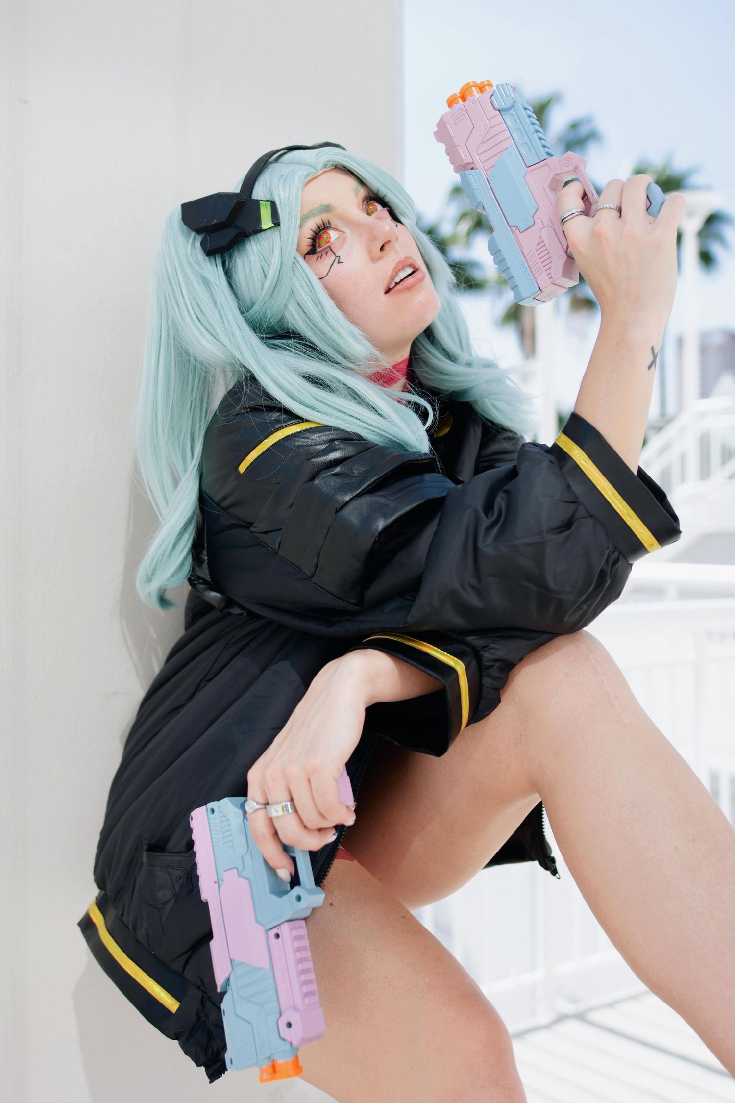 Cosplayer in black military-style uniform with mint green hair holds a toy gun against bright background.