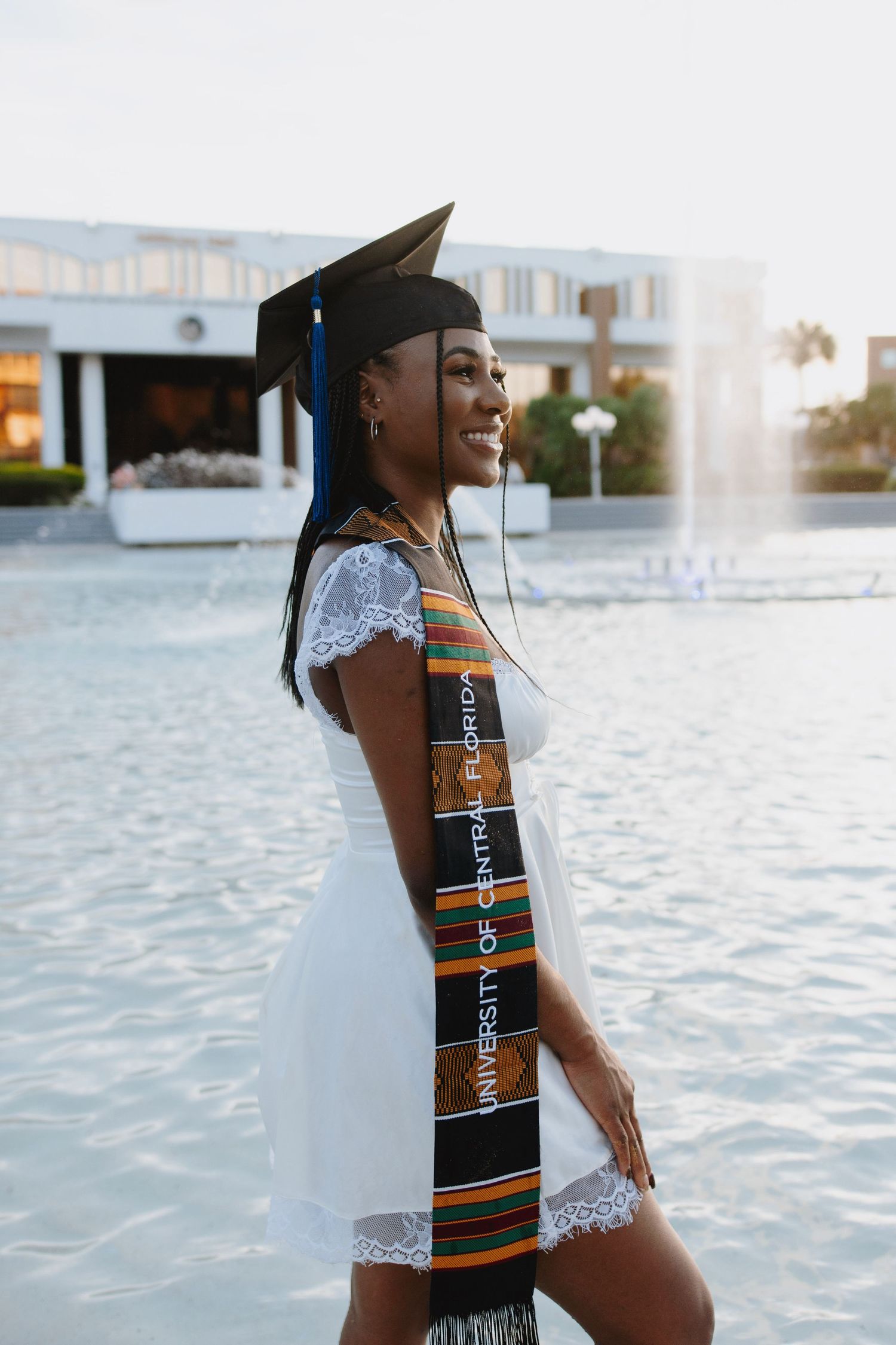 A graduate wearing a colorful graduation stole poses confidently by water fountain.