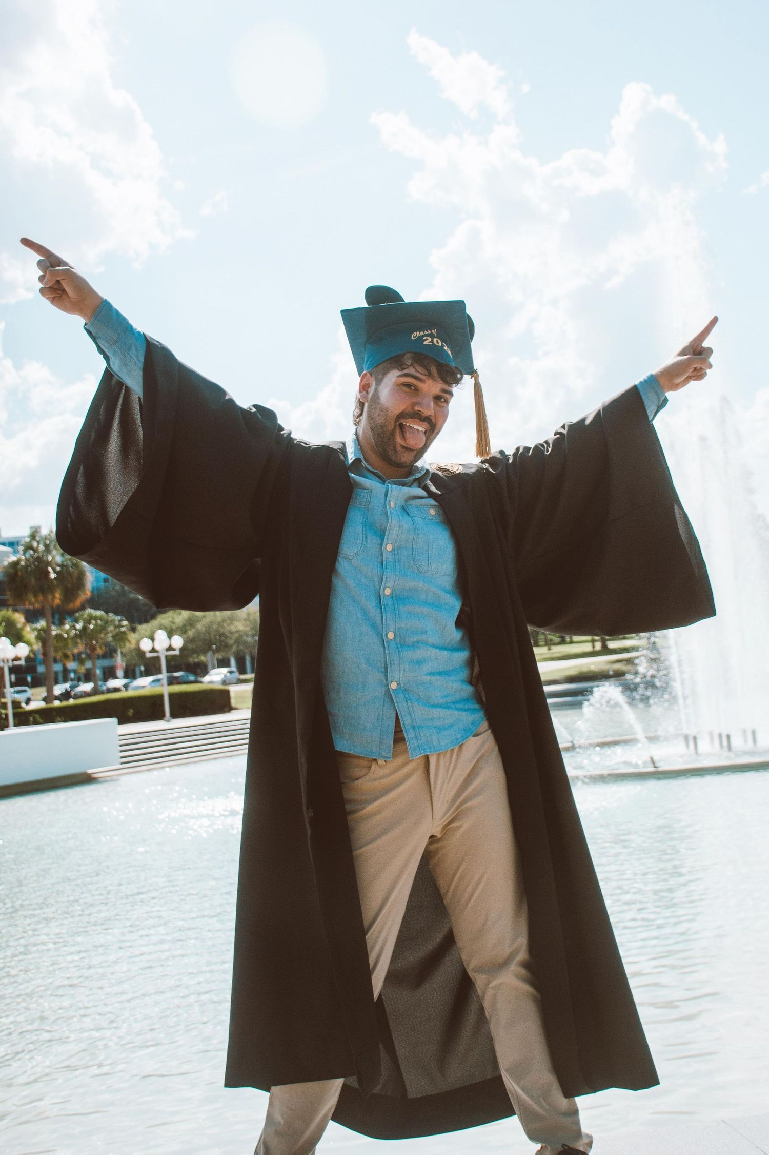 Graduate in academic regalia poses energetically by waterfront fountain.