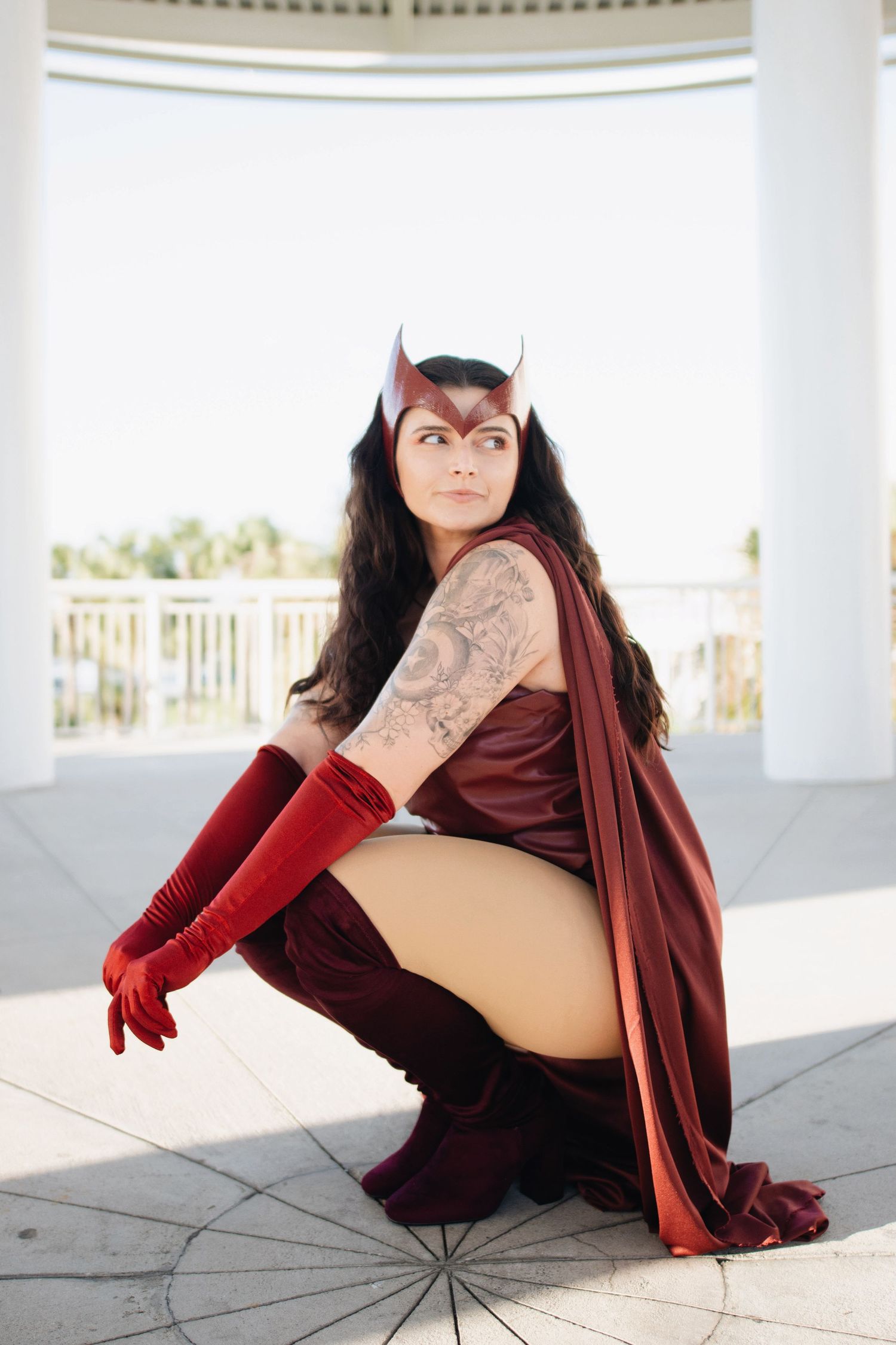 Cosplayer in maroon bodysuit and cape poses dramatically on white balcony with scenic background.