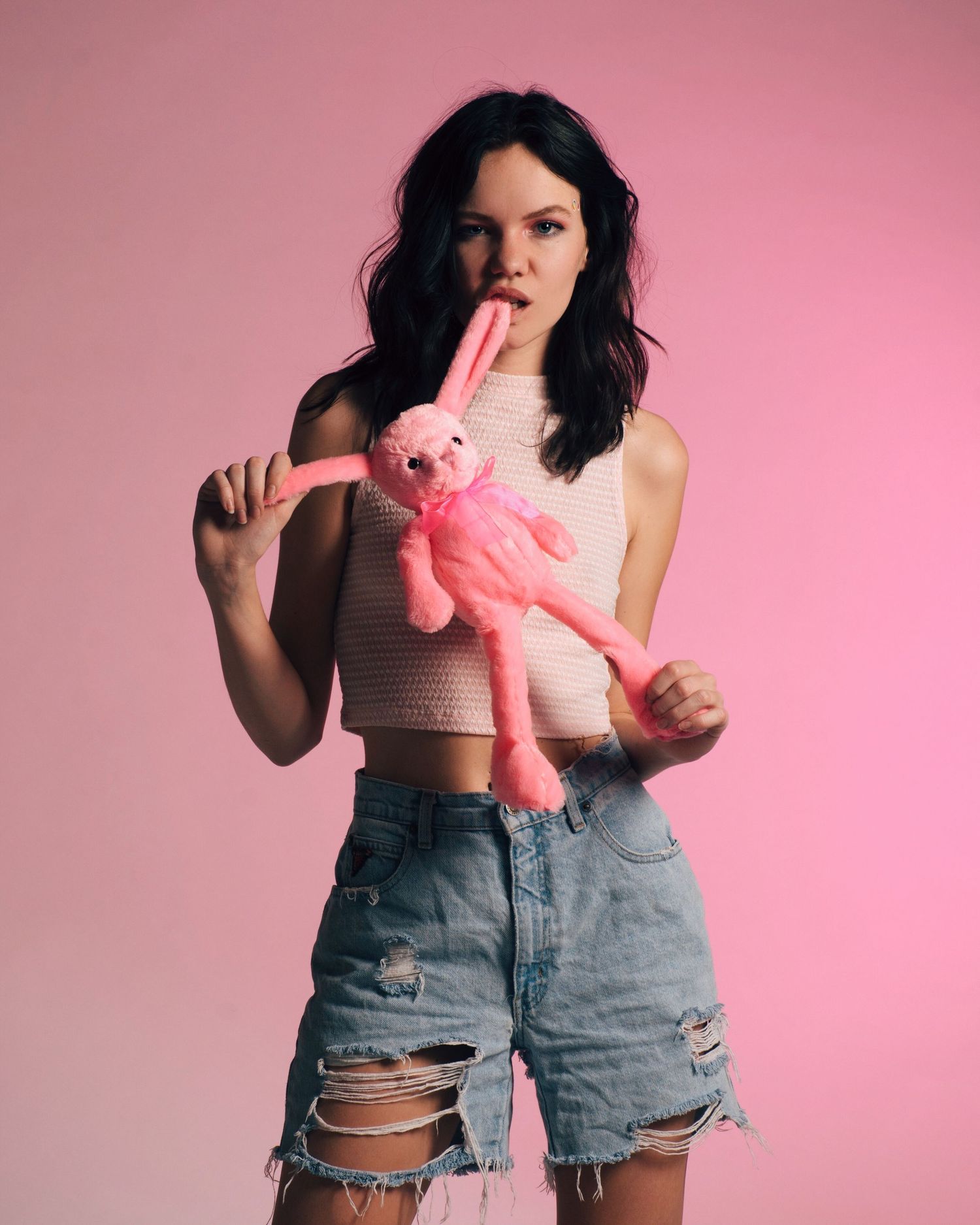 Playful fashion shoot with distressed denim shorts and pink styling against rose background.